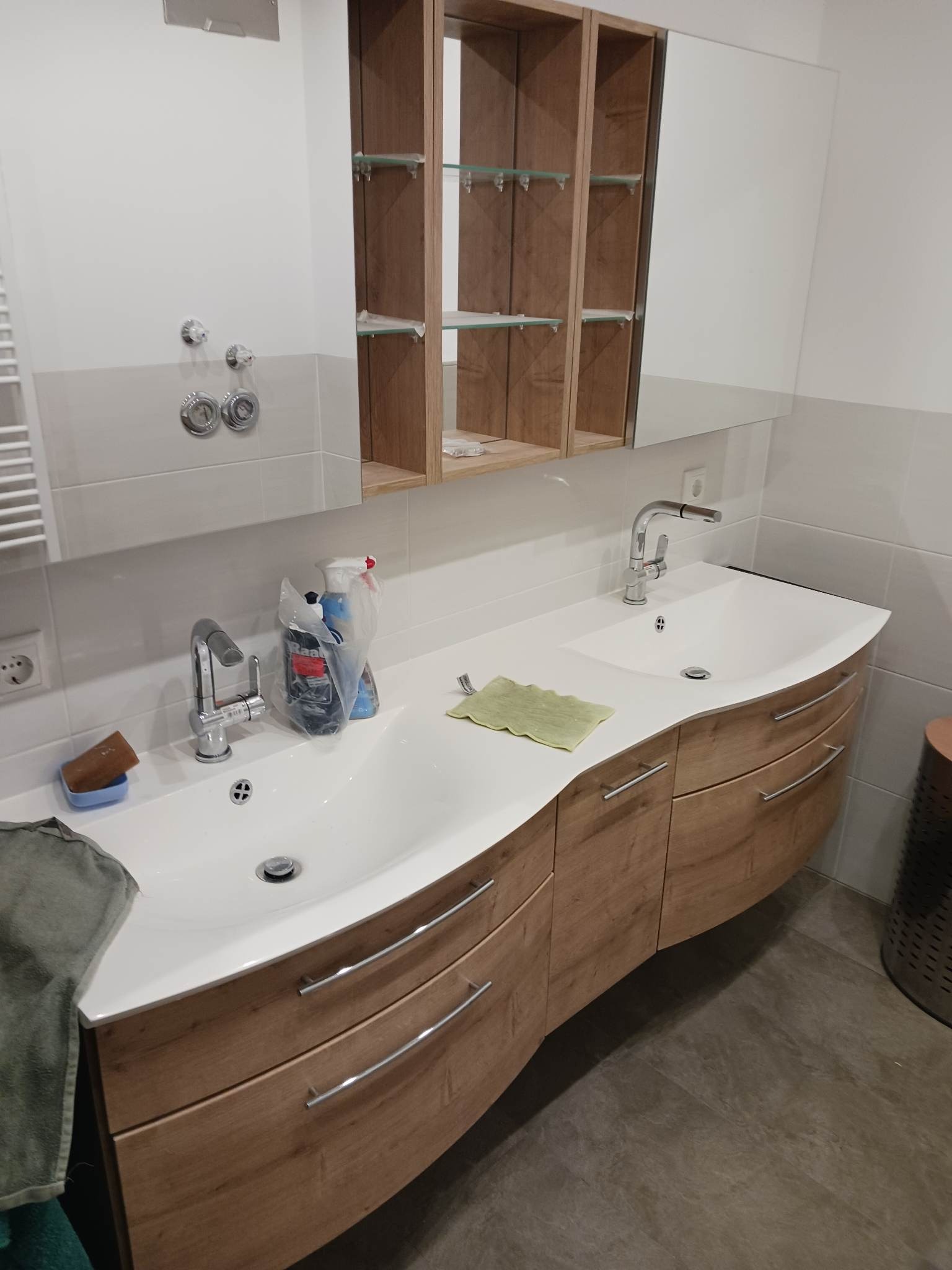 Nowoczesna łazienka z podwójną umywalką wpuszczoną w drewnianą szafkę z szufladami, nad którą wisi lustro z drewnianymi półkami. Na blacie umywalki leży zielona ściereczka i stoją środki czystości.