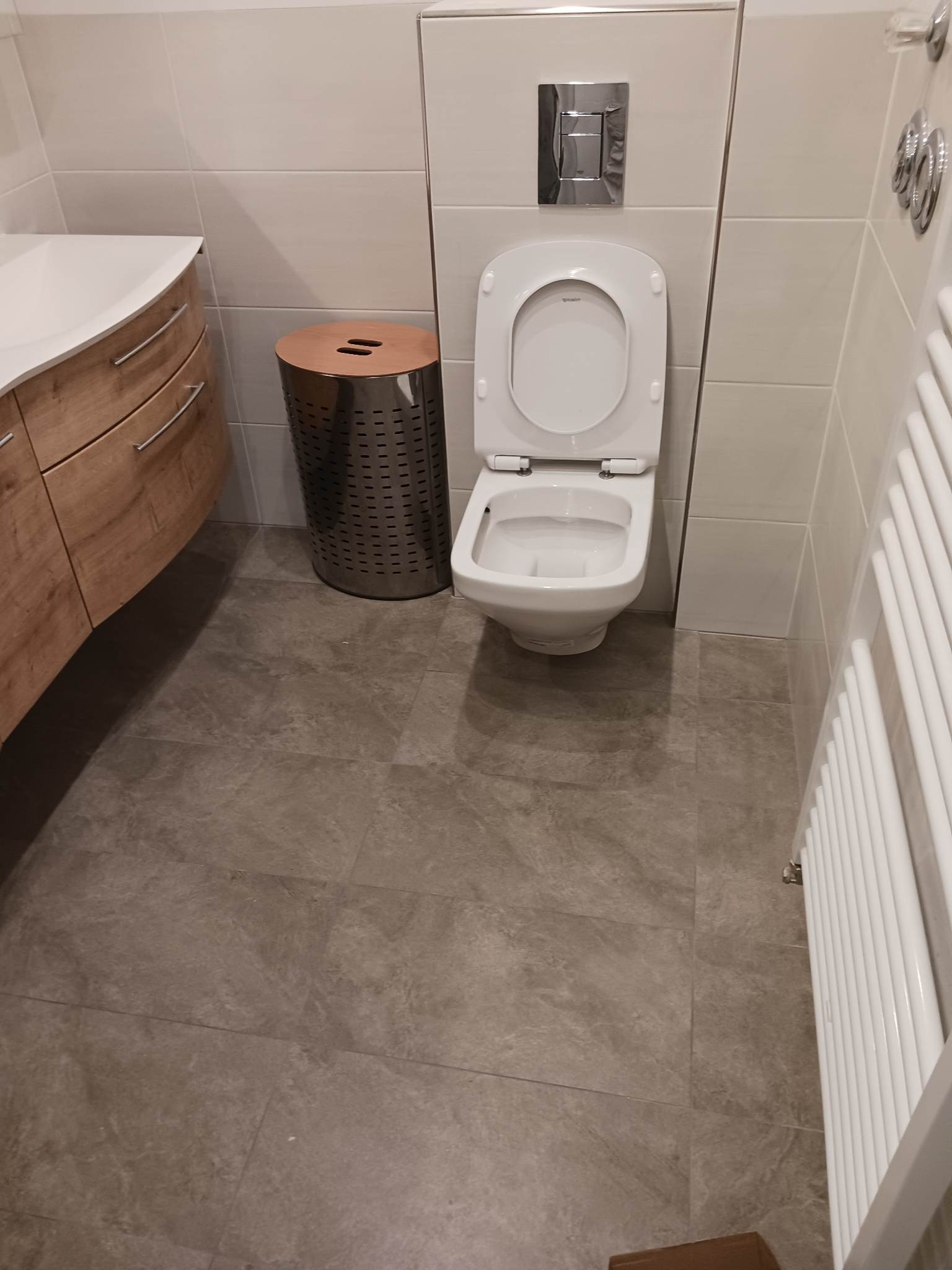 Nowoczesna łazienka z wiszącą toaletą Duravit, chromowanym przyciskiem spłukującym, grzejnikiem drabinkowym, szafką pod umywalkę z drewnopodobnym fornirem i metalowym koszem na pranie.