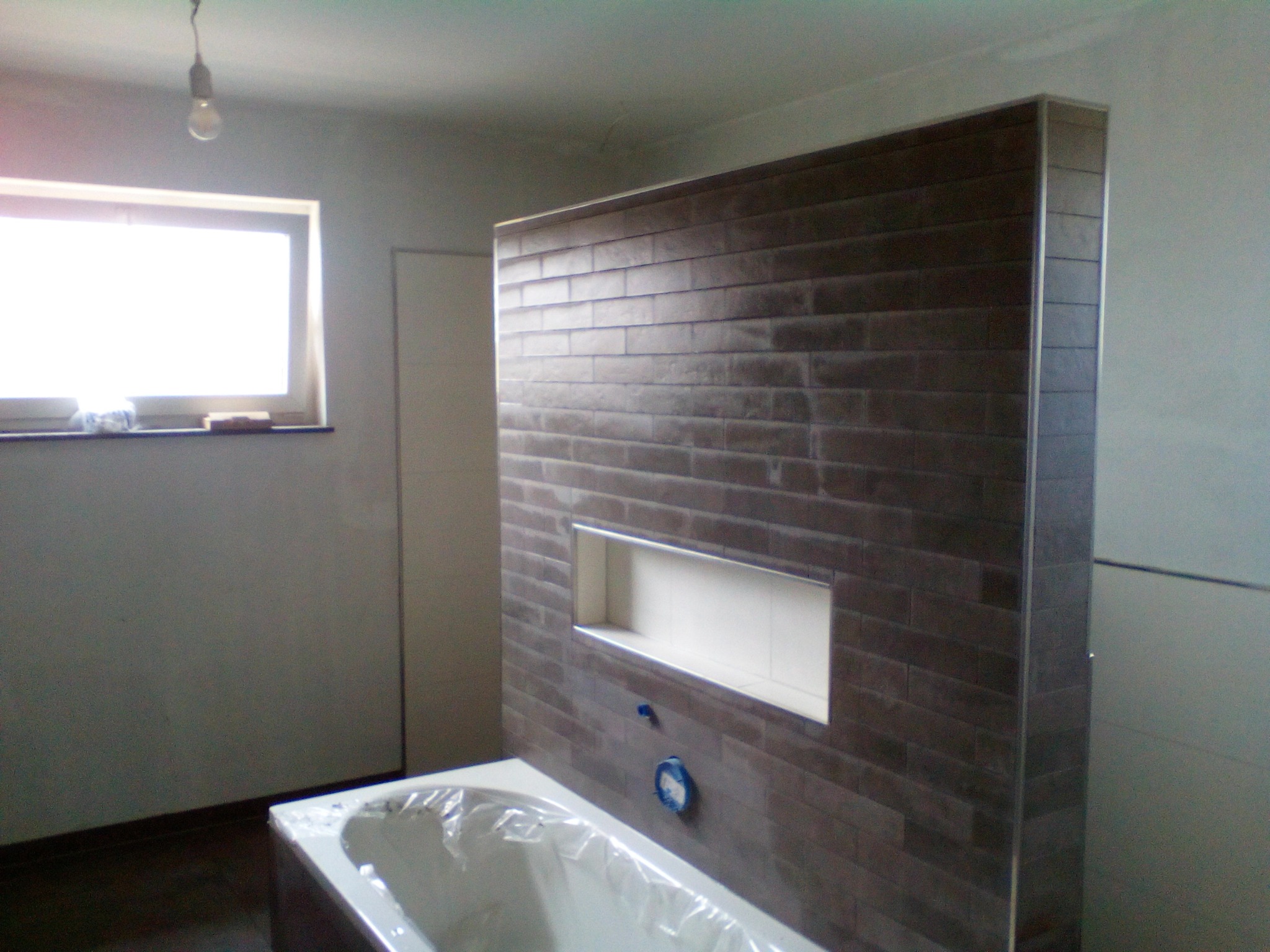 Częściowo wykończona łazienka z ciemnobrązową ścianą z płytek, wnęką, nową wanną zabezpieczoną folią i wiszącą żarówką.