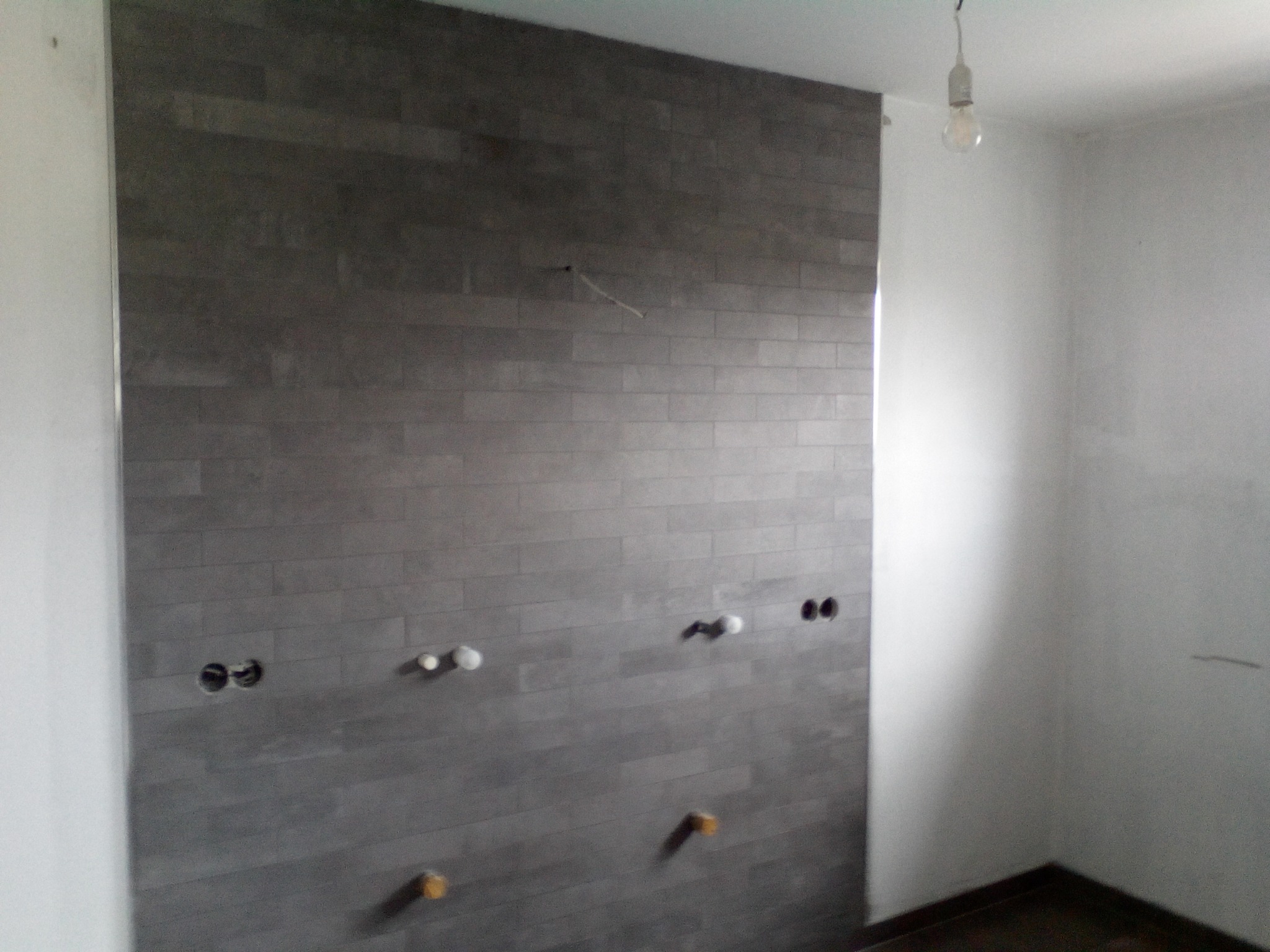 Ściana w łazience we Włocławku wyłożona szarymi płytkami imitującymi cegłę, z widocznymi wyprowadzeniami hydraulicznymi i elektrycznymi, surowa żarówka zwisa z sufitu.