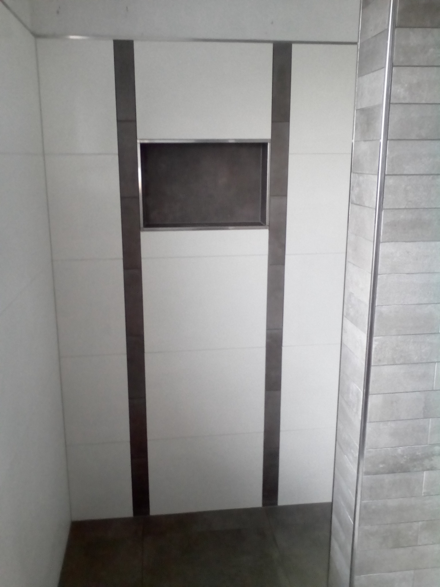 Wykończona ściana w łazience z białymi płytkami, ciemnymi pasami dekoracyjnymi i wnęką, widoczne także szare płytki na sąsiedniej ścianie.