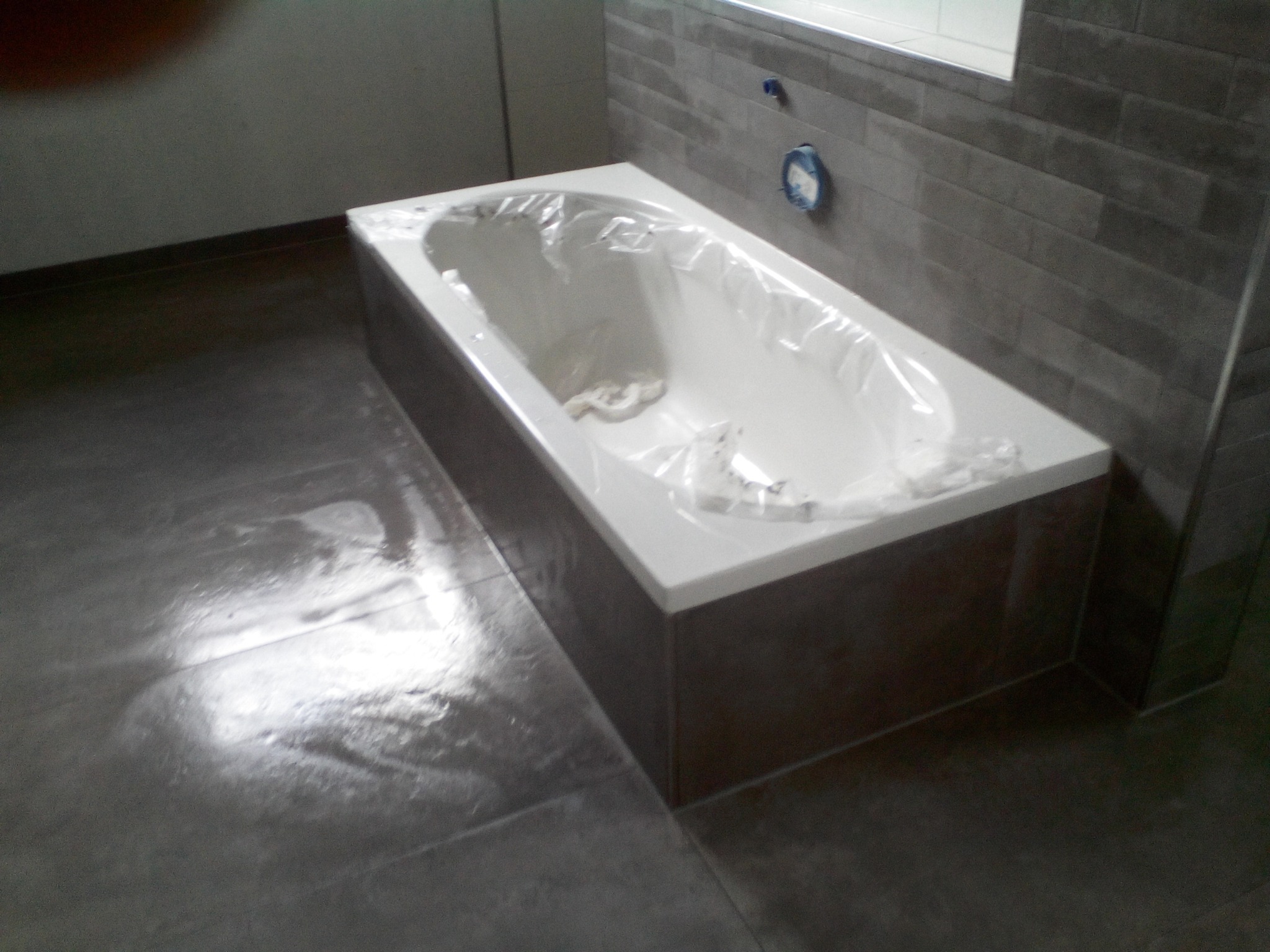 Nowa, biała wanna zabezpieczona folią ochronną w trakcie prac wykończeniowych w łazience, widoczne szare płytki na ścianach i podłodze, z pozostawionymi otworami na armaturę.