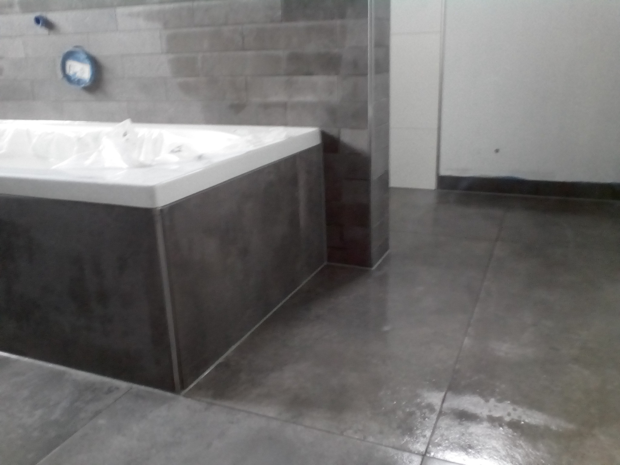 Nowoczesna łazienka w trakcie wykańczania: szare płytki na ścianach i podłodze, obudowana wanna z białą obudową, widoczne elementy instalacji.