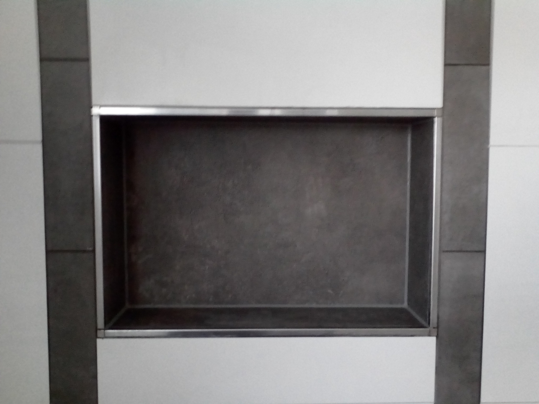 Wnęka w ścianie wyłożona ciemnoszarą płytką, obramowana srebrną listwą, otoczona białymi płytkami i pionowymi pasami z ciemnoszarej płytki.