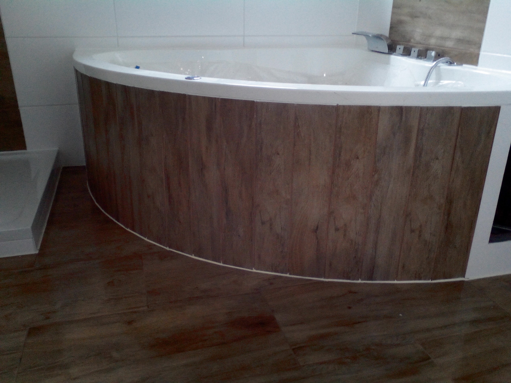 Zaokrąglona wanna z hydromasażem obudowana panelami drewnopodobnymi, widoczna z niskiego kąta na tle białych płytek i drewnianej podłogi.