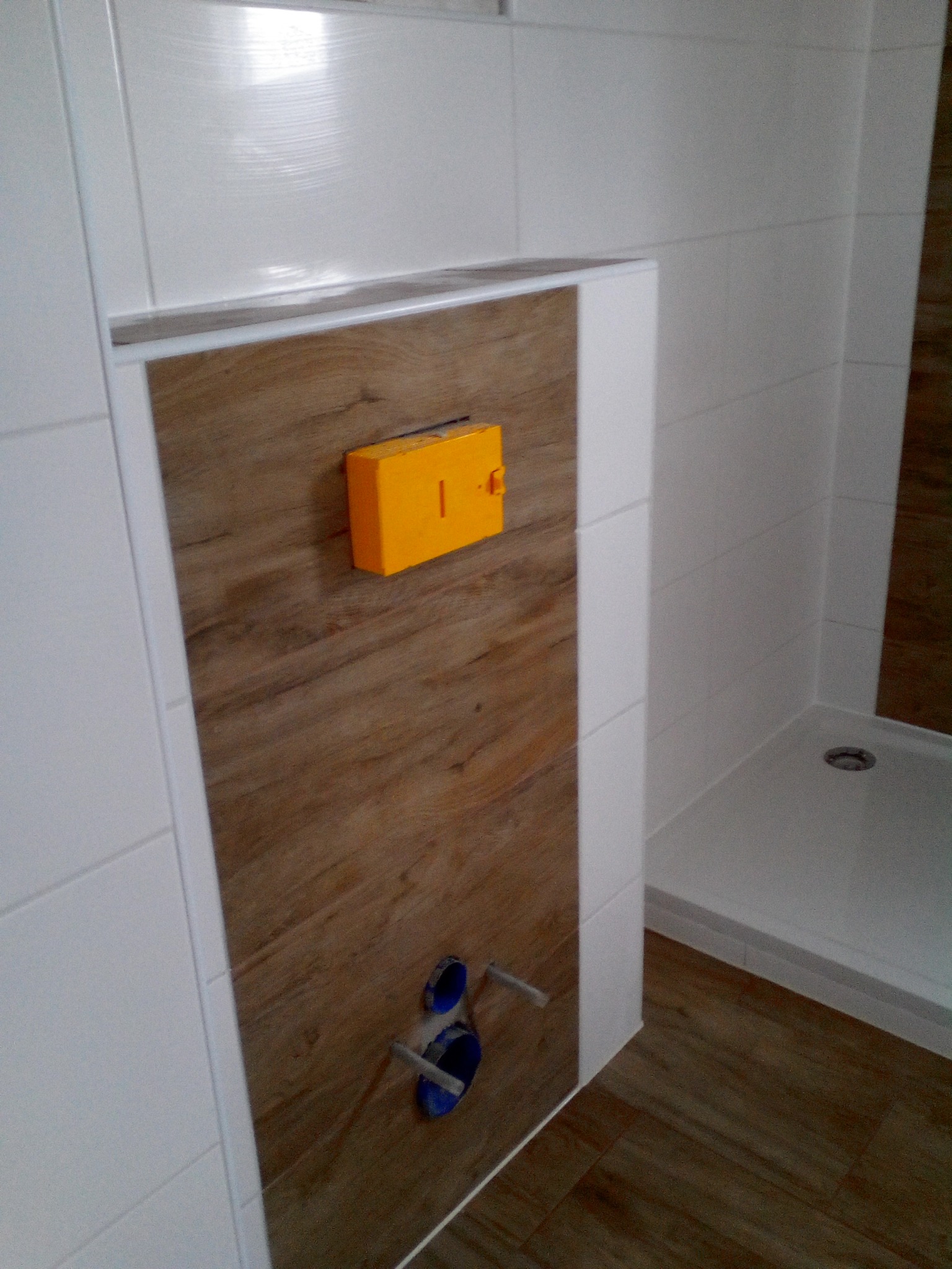 Fragment łazienki z zabudową stelaża WC wykończoną płytkami drewnopodobnymi, widoczne białe płytki na ścianach, żółty element instalacyjny i fragment kabiny prysznicowej.