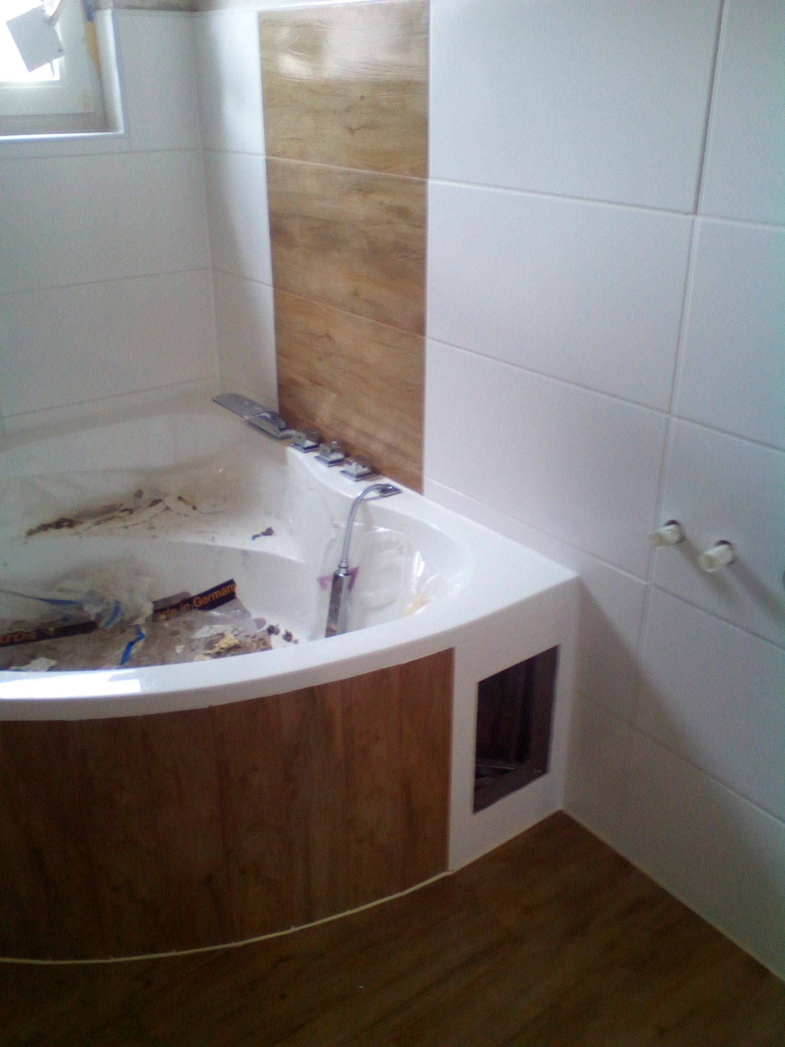 Narożna wanna z hydromasażem w trakcie montażu, widoczne białe płytki ścienne z dekoracyjnym pasem imitującym drewno, niedokończona zabudowa wanny z drewnopodobną okleiną, podłoga również z paneli...