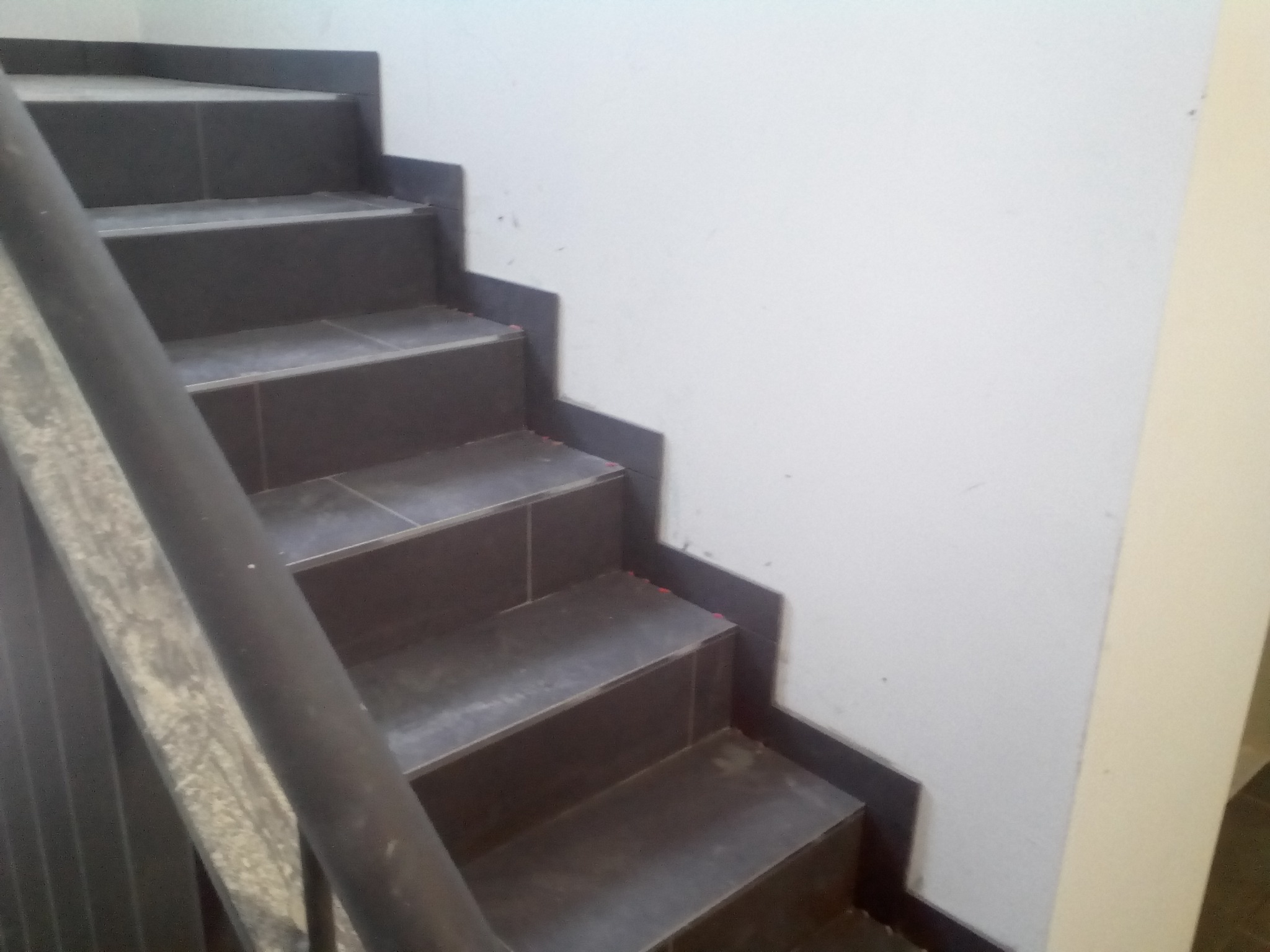 Schody wyłożone ciemnymi płytkami, z metalową poręczą i białą ścianą w tle. Widoczne drobne niedociągnięcia w wykończeniu.