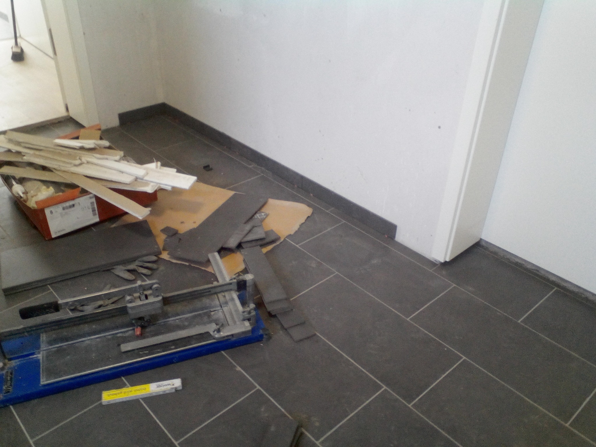 Fragment podłogi z ułożonymi ciemnymi płytkami, obok leżą płytki, skrzynka z resztkami, gąbka i przecinarka do płytek.