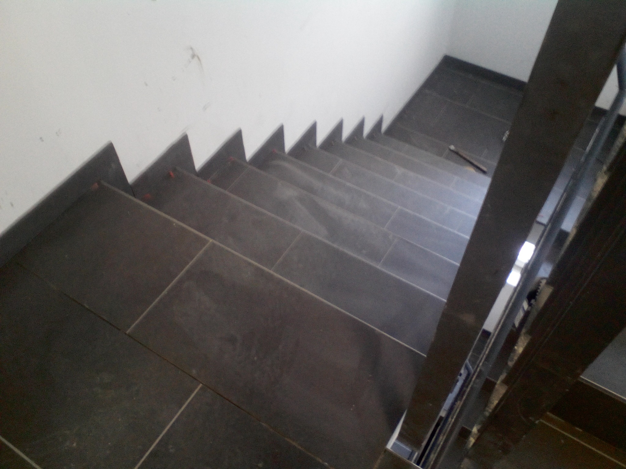 Schody wyłożone ciemnymi, prostokątnymi płytkami, widok z góry, fragment metalowej barierki po prawej stronie, ściana biała, na schodach leży ołówek.