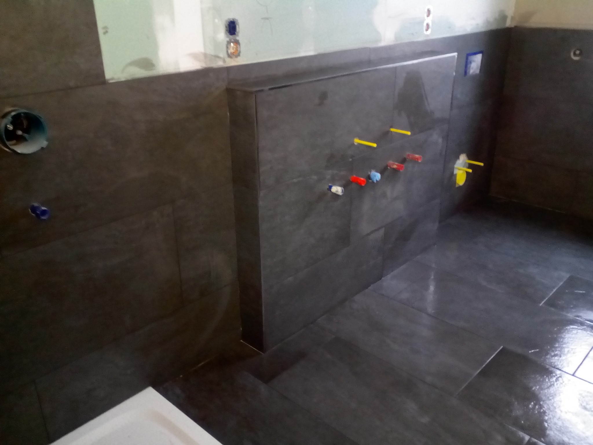 Ciemne, płytki imitujące drewno na ścianach i podłodze w trakcie prac. Widoczne przyłącza wodne i elektryczne, oraz fragment białego elementu wyposażenia.