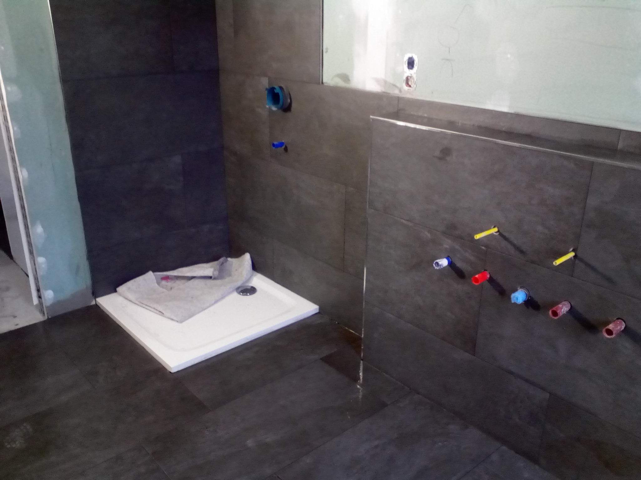 Montaż brodzika w łazience z ciemnymi płytkami imitującymi drewno na ścianach i podłodze; widoczne wystające rury instalacji wodnej z kolorowymi zaślepkami.