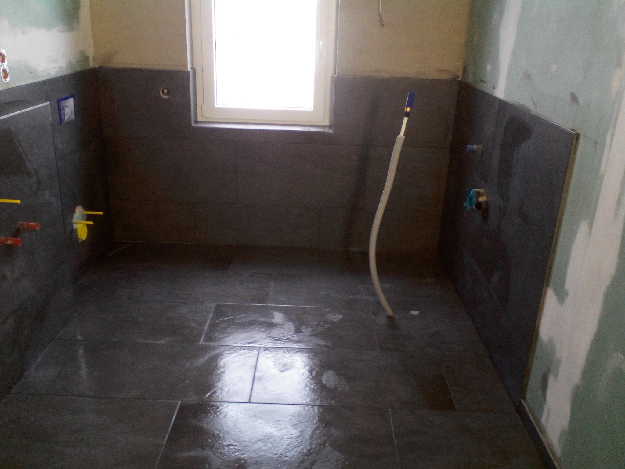 Wnętrze łazienki w trakcie prac wykończeniowych: ułożone ciemne płytki na podłodze i ścianach do połowy wysokości, widoczne rury instalacji wodnej i okno z białymi ramami.