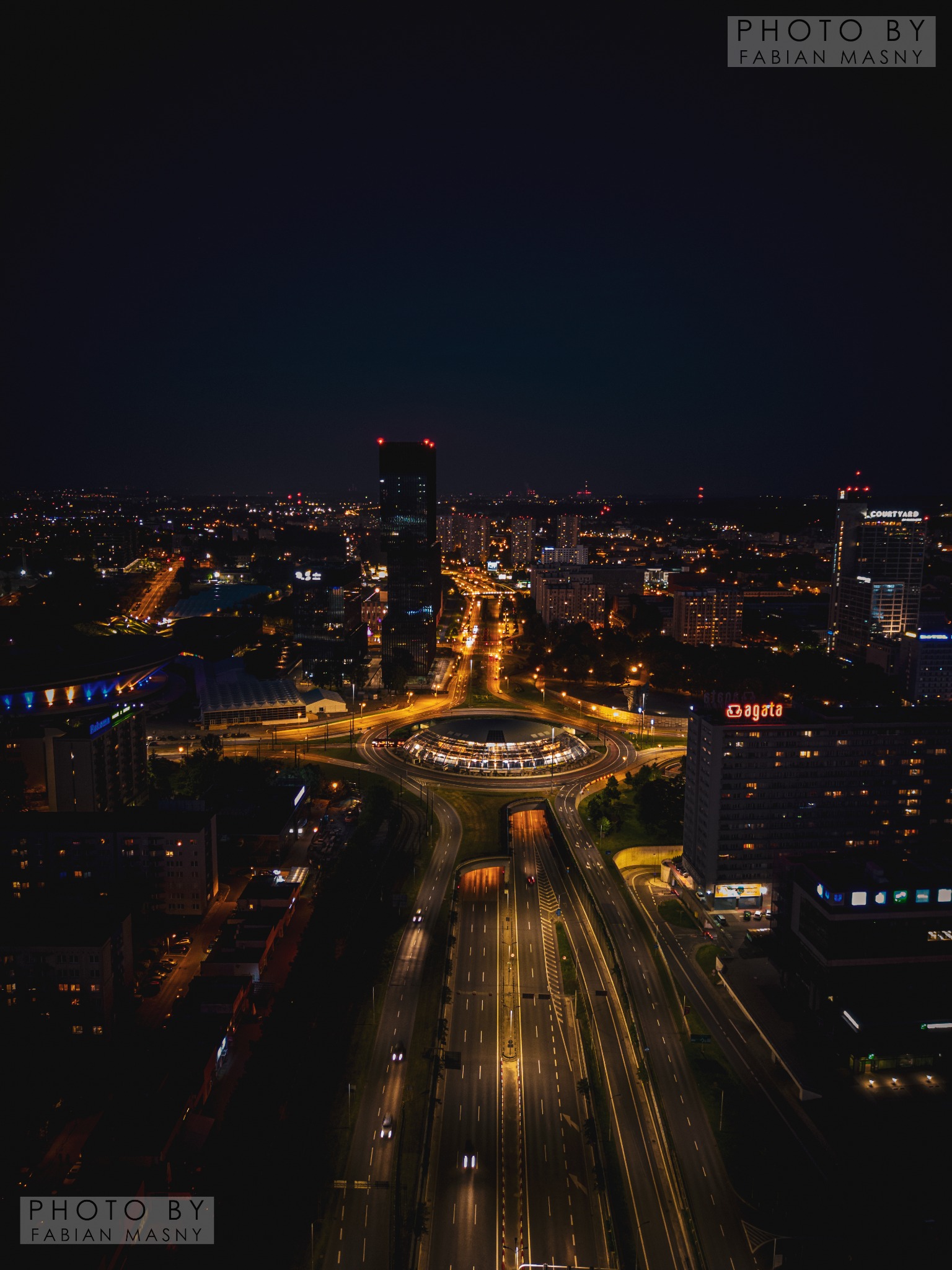 Nocna panorama miasta z wysokiego punktu widzenia, ukazująca oświetlone ulice, ronda i budynki, w tym charakterystyczny wieżowiec z czerwonymi światłami na szczycie oraz hotel Courtyard.