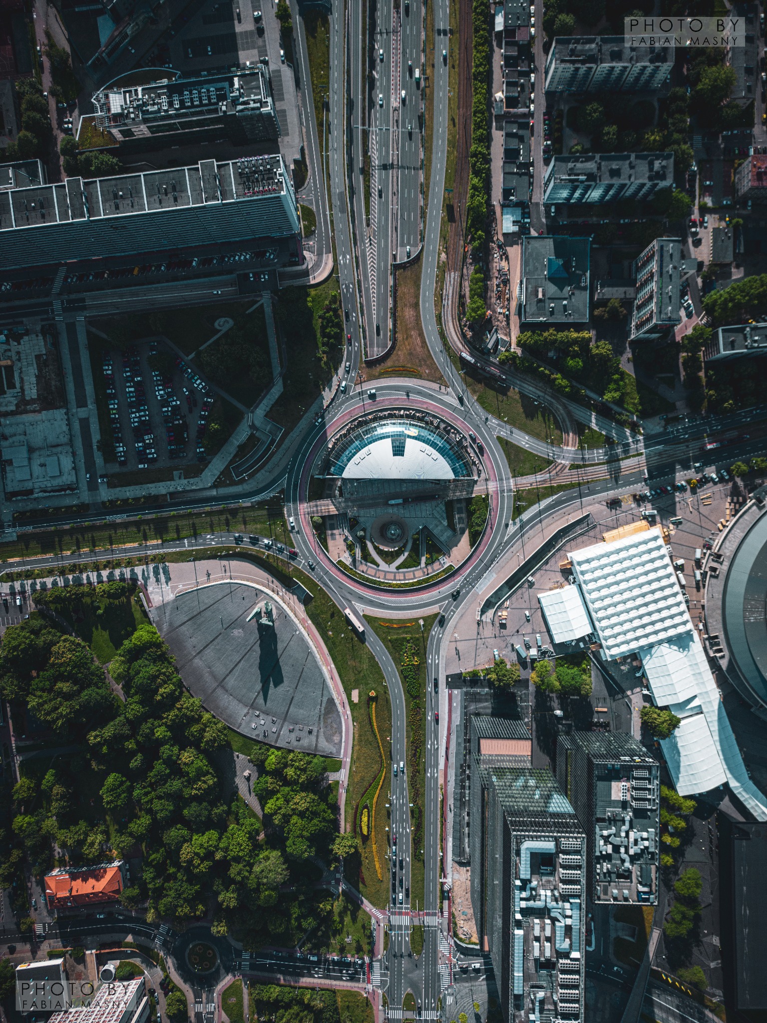 Widok z drona na rondo z charakterystycznym budynkiem pośrodku, otoczone zielenią, drogami i budynkami, z widocznymi samochodami i pieszymi, w słoneczny dzień.