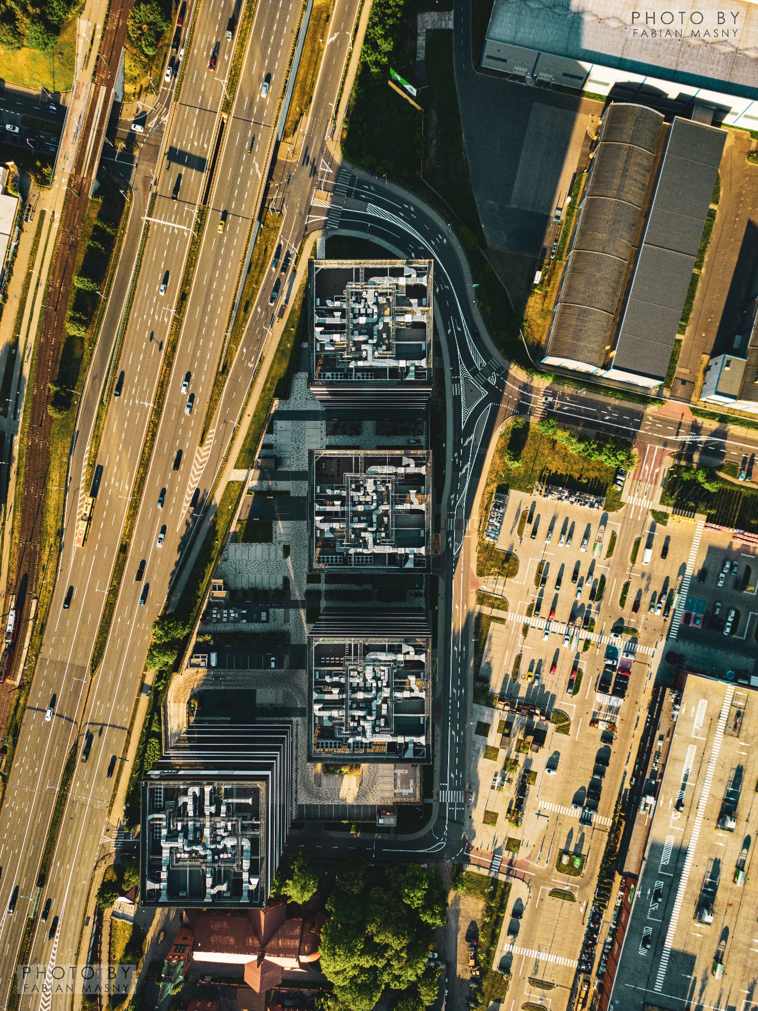 Zdjęcie z drona przedstawia kompleks biurowy z charakterystycznymi instalacjami na dachach, przylegający do ruchliwej autostrady i rozległego parkingu.