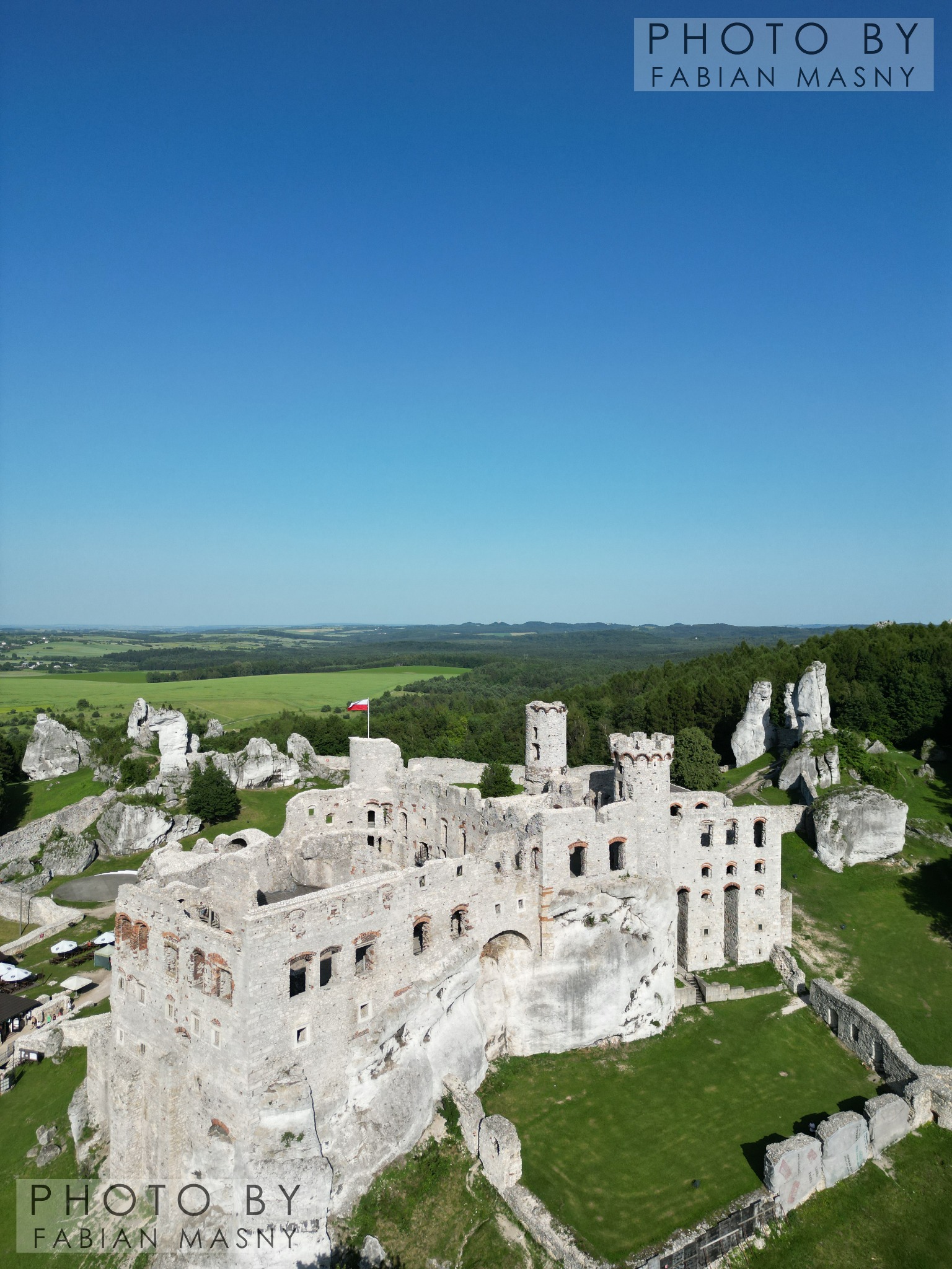 Widok z lotu ptaka na ruiny zamku Ogrodzieniec z powiewającą polską flagą, wapienne skały i zielone wzgórza w tle pod błękitnym niebem.