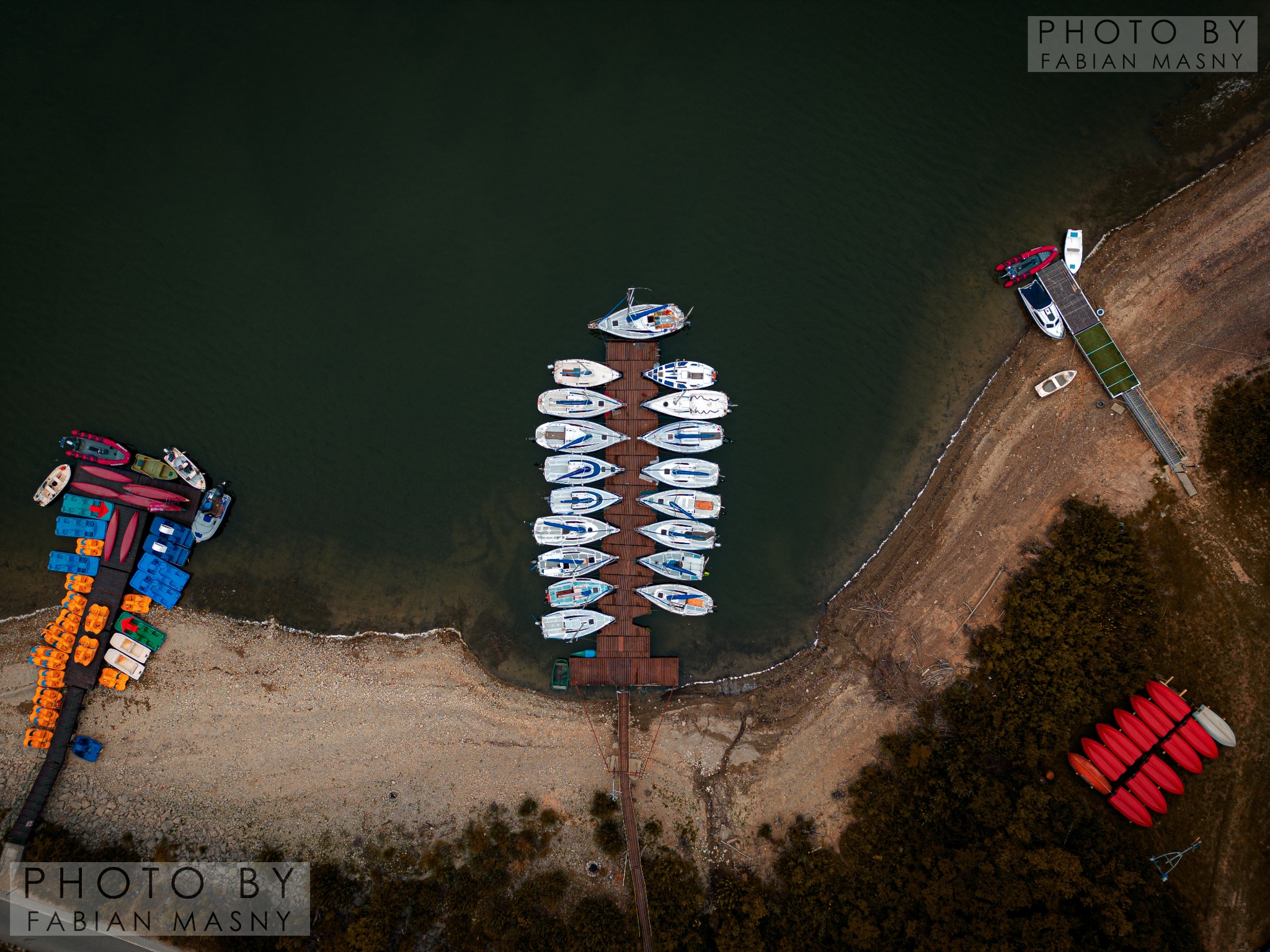 Widok z lotu ptaka na przystań z rzędem jachtów, kajaki na brzegu i pomost z łodziami, otoczone brązową linią brzegową i ciemną wodą.