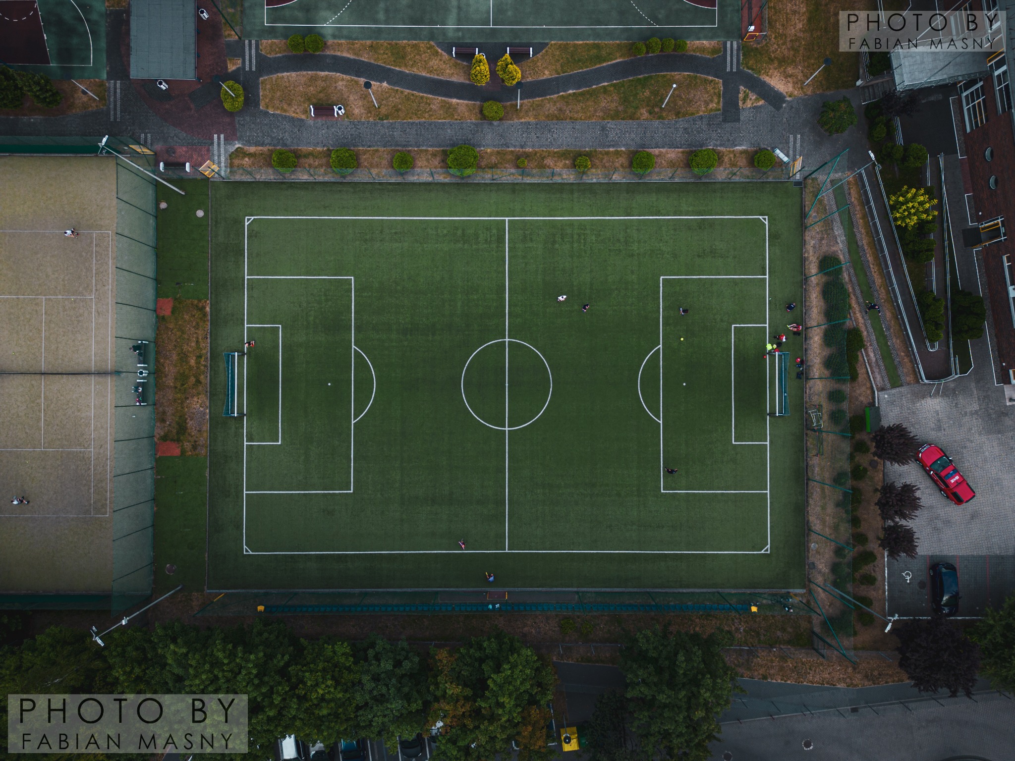 Widok z drona na boisko piłkarskie z zieloną murawą i białymi liniami, otoczone innymi boiskami, ścieżkami i drzewami. Na boisku widoczni piłkarze podczas treningu.