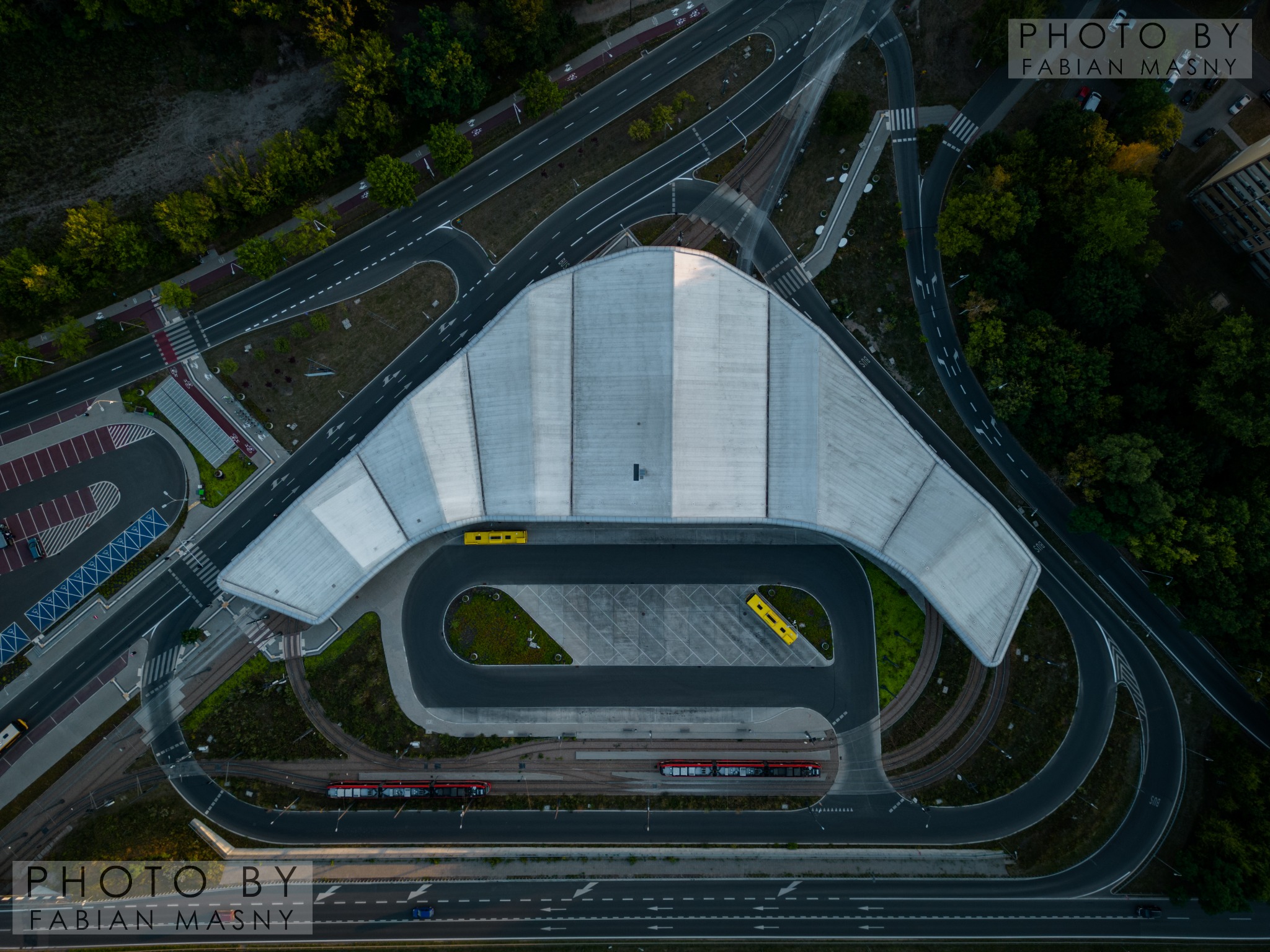 Zdjęcie z drona zajezdni autobusowej i tramwajowej z charakterystycznym dachem w kształcie litery V, otoczonej drogami i zielenią, z widocznymi autobusami i tramwajami.
