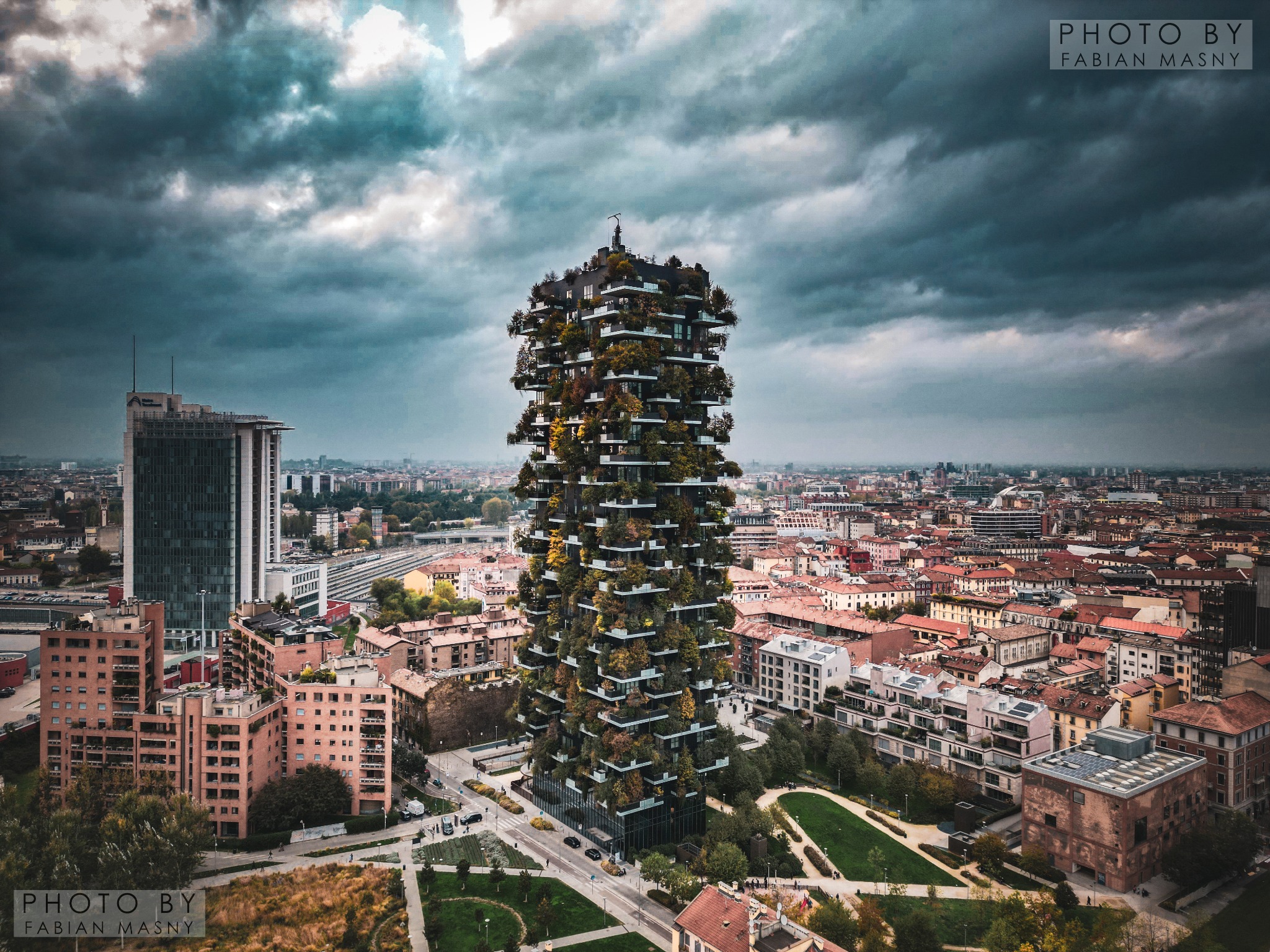 Wysoki budynek mieszkalny porośnięty zielenią, na tle panoramy miasta i pochmurnego nieba.