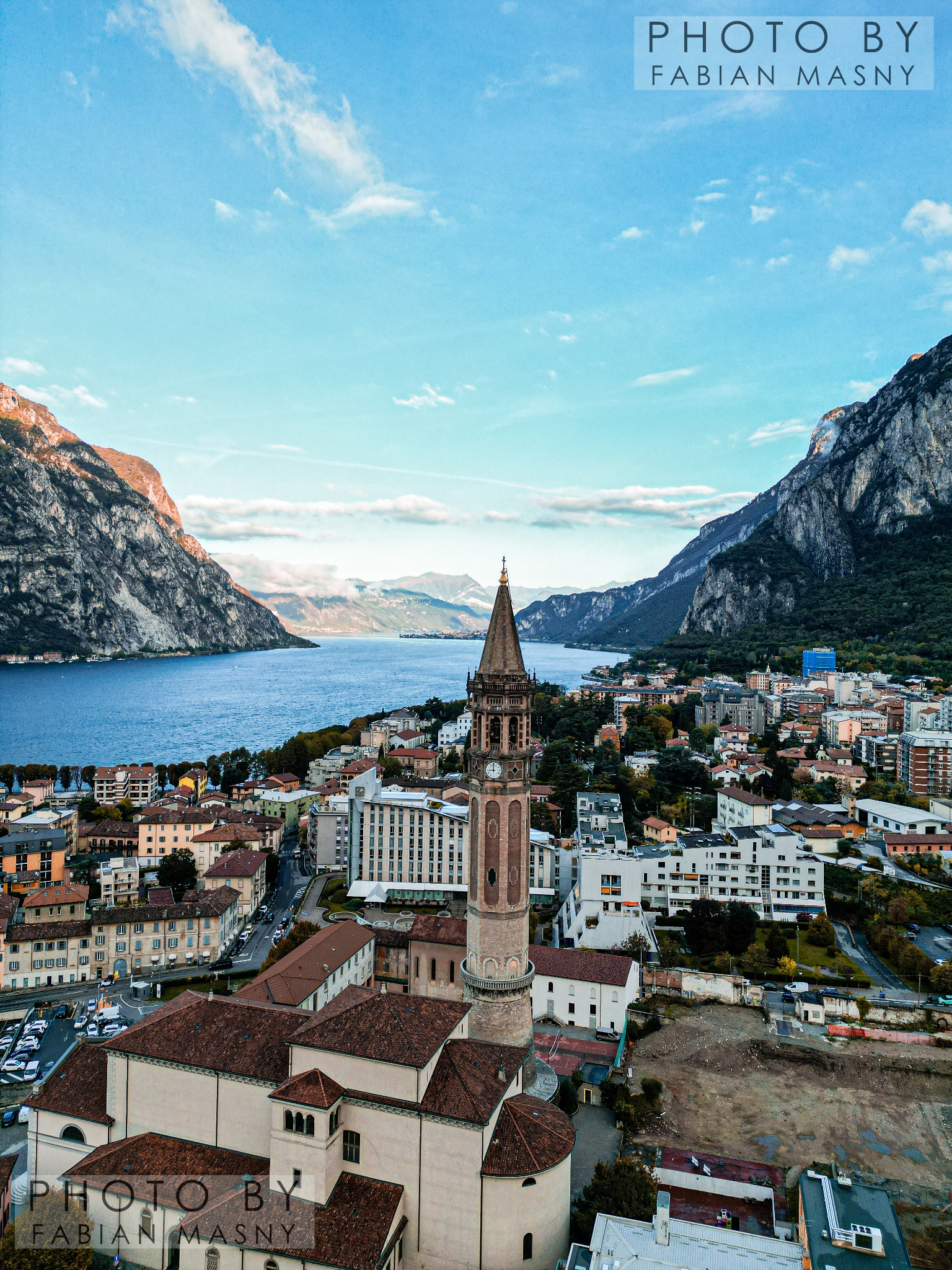 Panorama miasta Lecco we Włoszech z widokiem na jezioro Como, otoczone górami, z wieżą kościoła w pierwszym planie, sfotografowane z góry.
