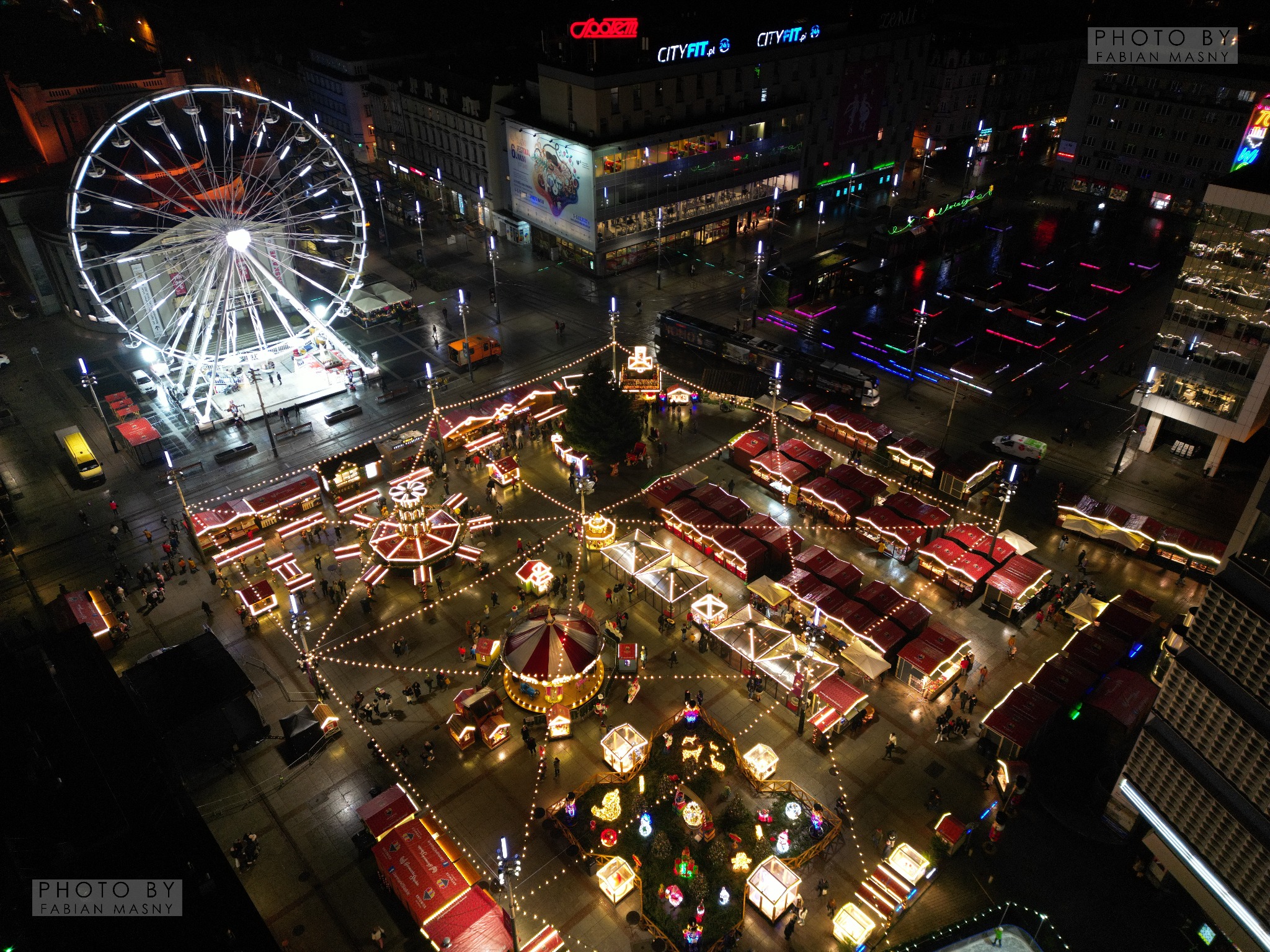 Nocny widok z lotu ptaka na jarmark bożonarodzeniowy z karuzelą, straganami i oświetlonym diabelskim młynem w tle.