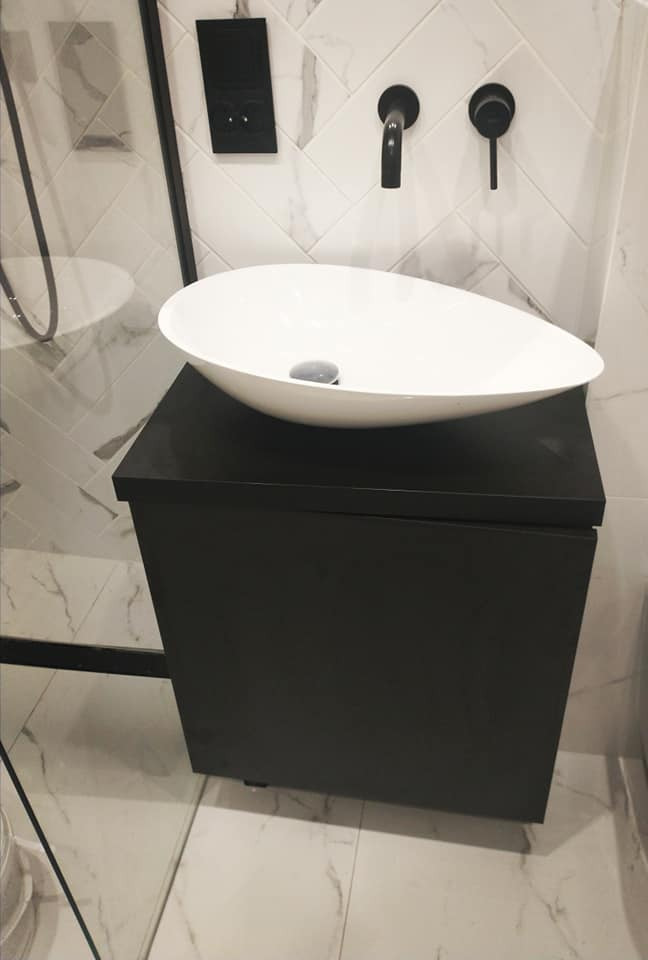 Biała, owalna umywalka nablatowa na czarnej szafce łazienkowej, z czarną armaturą na tle białych płytek w jodełkę.