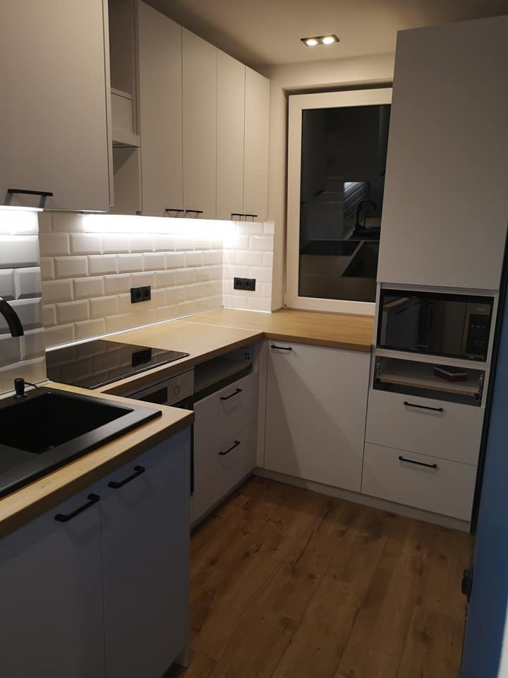 Nowoczesna kuchnia z szarymi frontami, drewnianym blatem, czarnym zlewozmywakiem i białymi płytkami typu metro podświetlonymi listwą LED.