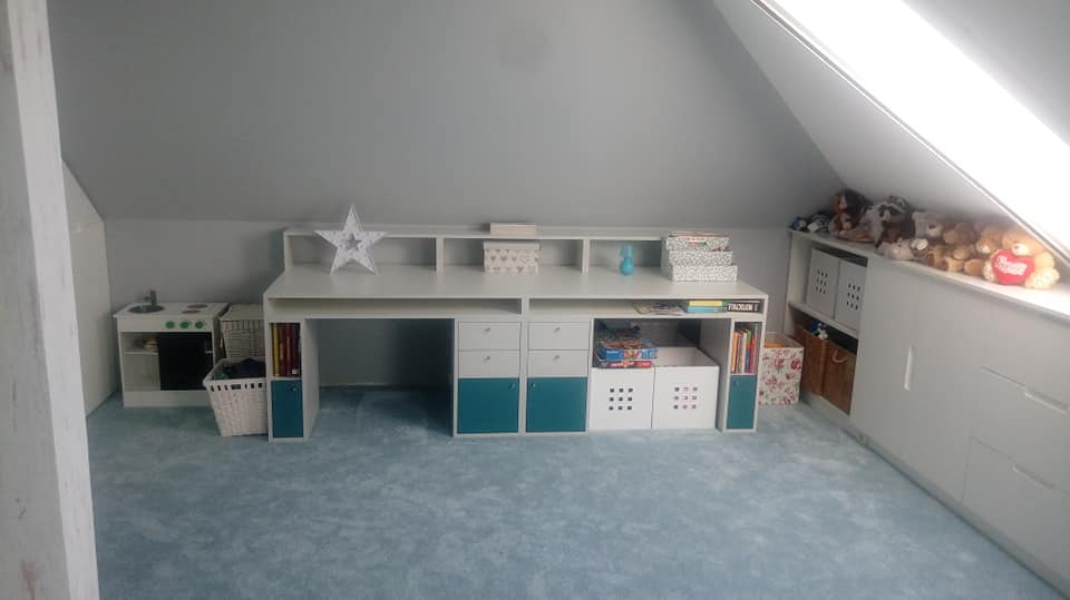 Białe biurko z nadstawką i szafkami w pokoju na poddaszu, z turkusowymi akcentami, obok biała kuchnia dla dzieci i półki z zabawkami