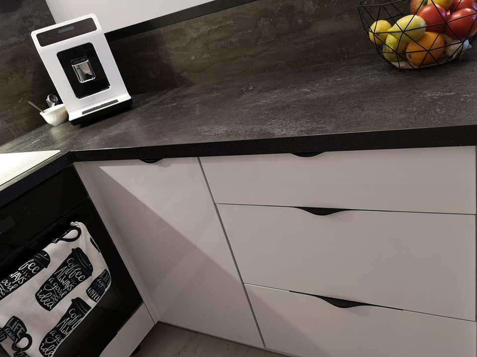 Fragment nowoczesnej kuchni z białymi, lakierowanymi frontami szafek i ciemnoszarym blatem z owocami w drucianym koszu, ekspres do kawy na blacie.