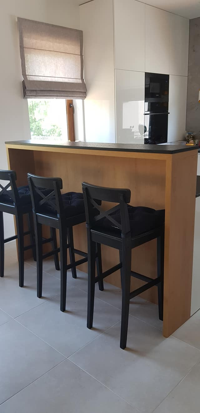 Nowoczesna kuchnia z drewnianą wyspą i czarnymi hokerami z tapicerowanymi siedziskami, minimalistyczne białe szafki w tle, roleta rzymska w oknie.