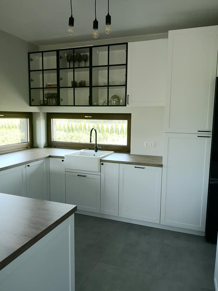 Jasna kuchnia z białymi szafkami i drewnianymi blatami, górne szafki przeszklone z czarnymi ramami, widok na zieleń za oknem, minimalistyczne lampy wiszące