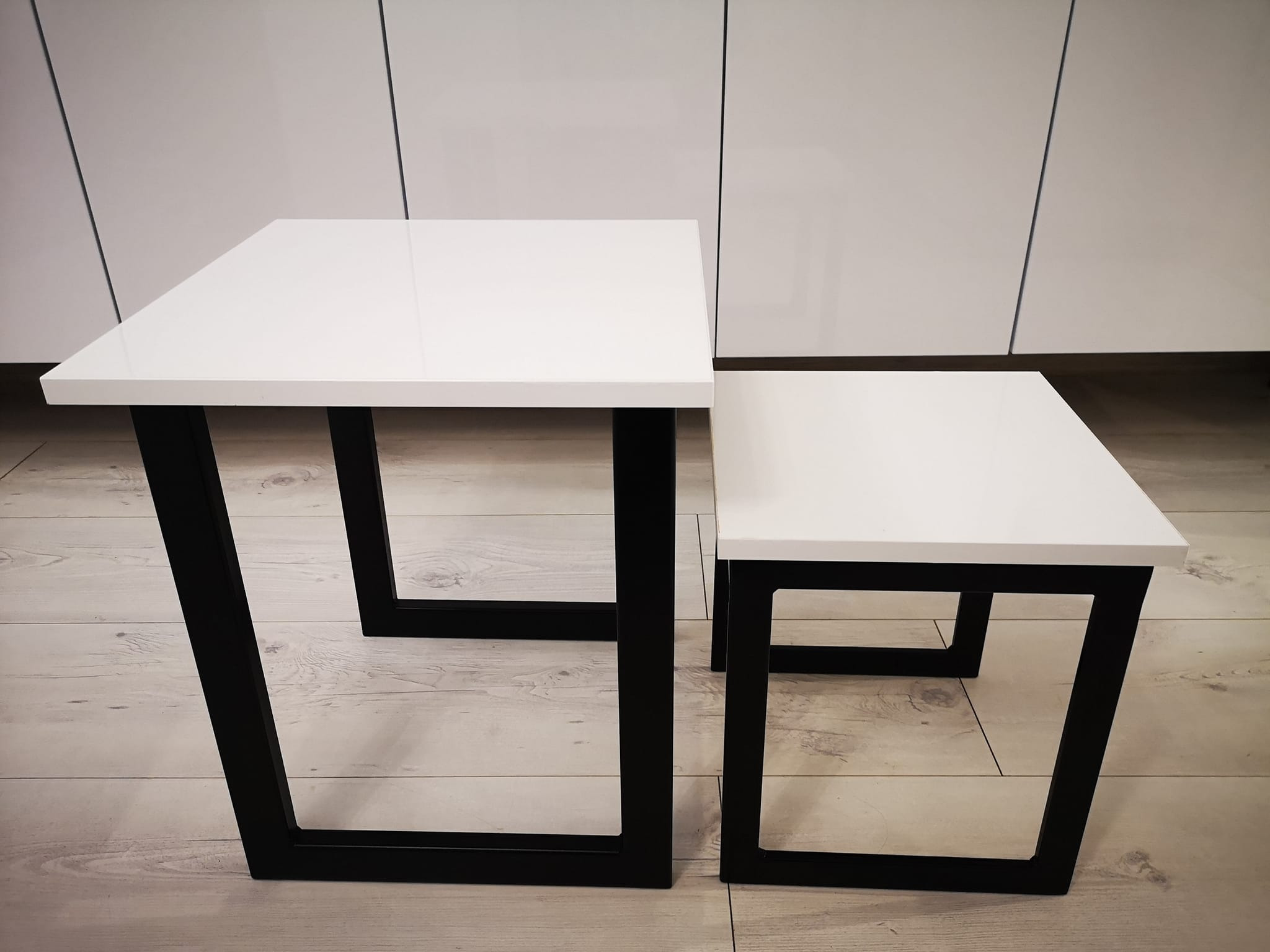 Dwa białe stoliki kawowe o kwadratowych blatach i czarnych, metalowych nogach, ustawione jeden obok drugiego na drewnianej podłodze, na tle białych szafek.