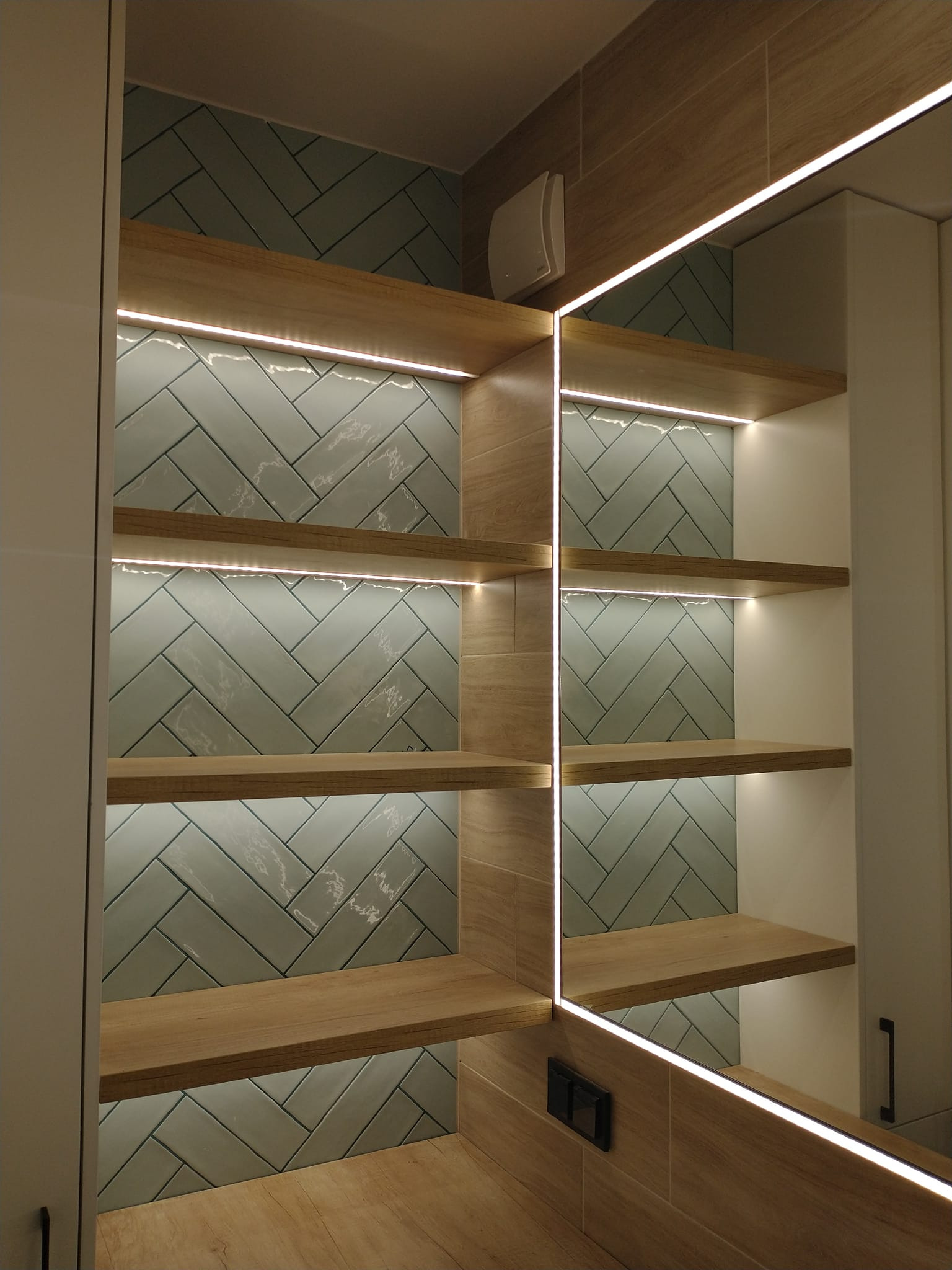 Półki drewniane z oświetleniem LED na tle ściany wyłożonej płytkami w jodełkę, widoczne odbicie w lustrze i fragment szafki.