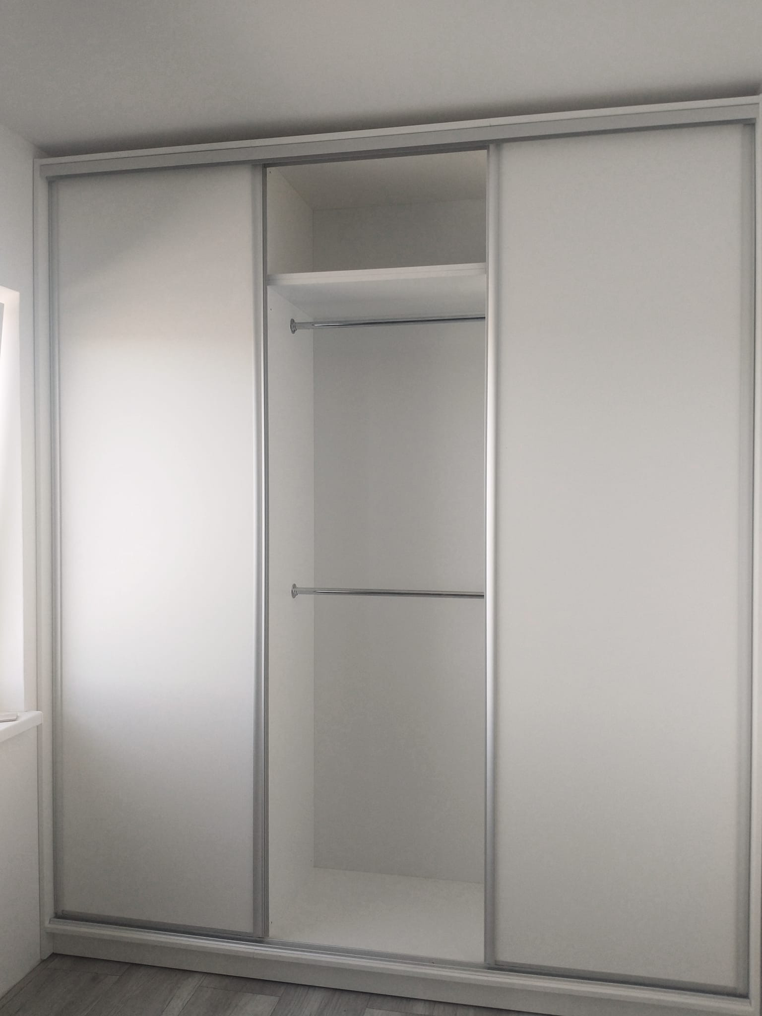 Biała szafa wnękowa z przesuwnymi drzwiami i metalowymi prowadnicami, widoczne półki i drążki na ubrania.