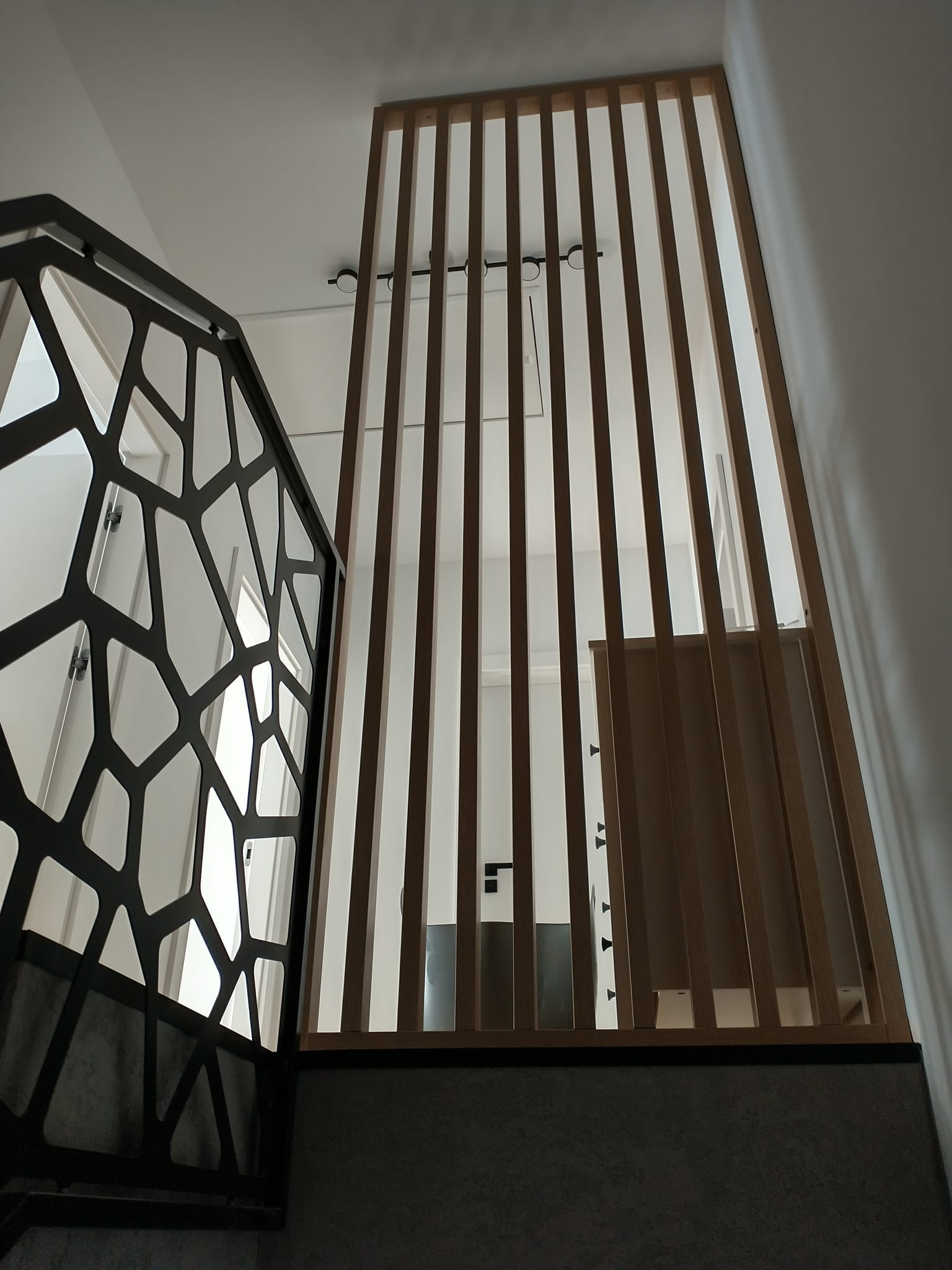 Ażurowa, metalowa balustrada schodowa z geometrycznym wzorem i drewniana ścianka działowa z pionowych listew w nowoczesnym wnętrzu, widok z dołu.