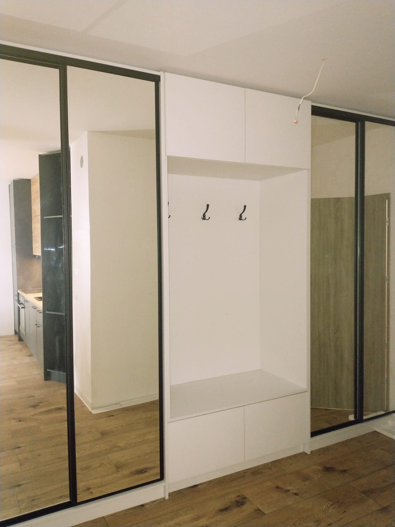 Biała zabudowa przedpokoju z siedziskiem, wieszakami i szafami przesuwnymi z lustrami w czarnych ramach, widok na kuchnię w tle.