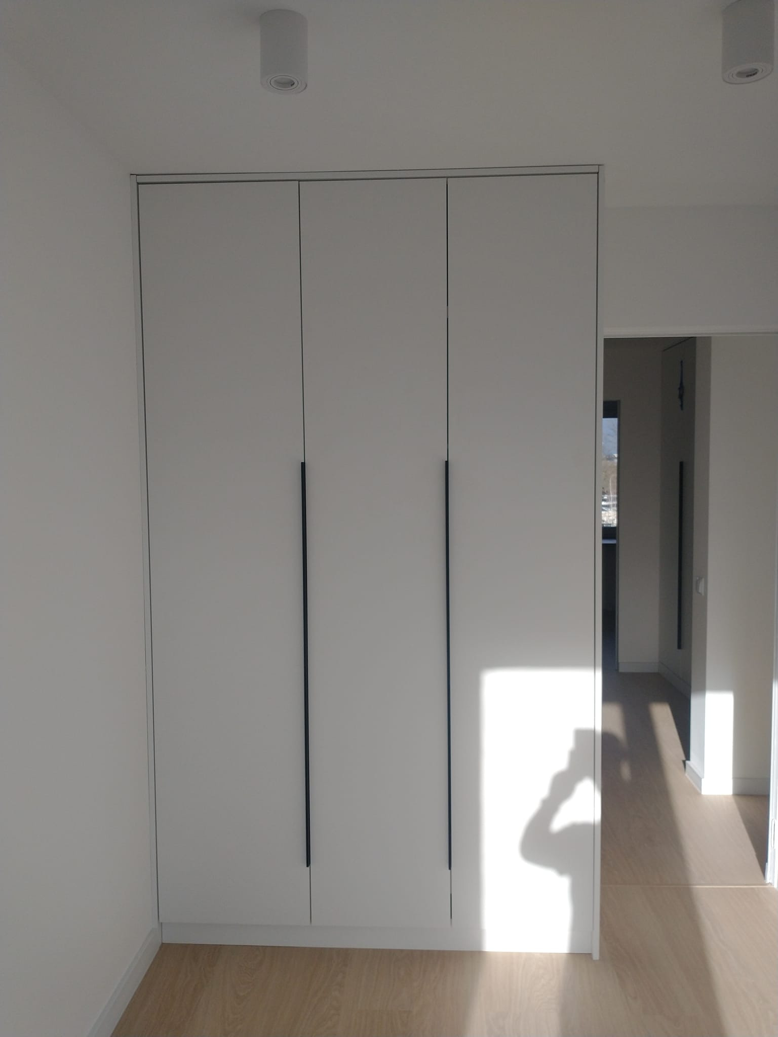 Trzydrzwiowa szafa wnękowa z białymi frontami i długimi, pionowymi uchwytami w minimalistycznym wnętrzu z parkietem, cień fotografa widoczny na szafie.