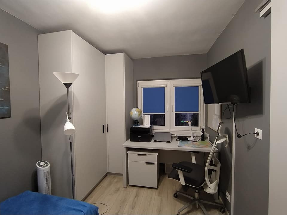 Szara sypialnia z biurkiem, telewizorem na ścianie i dużą białą szafą narożną, widok na nocne miasto przez okno.