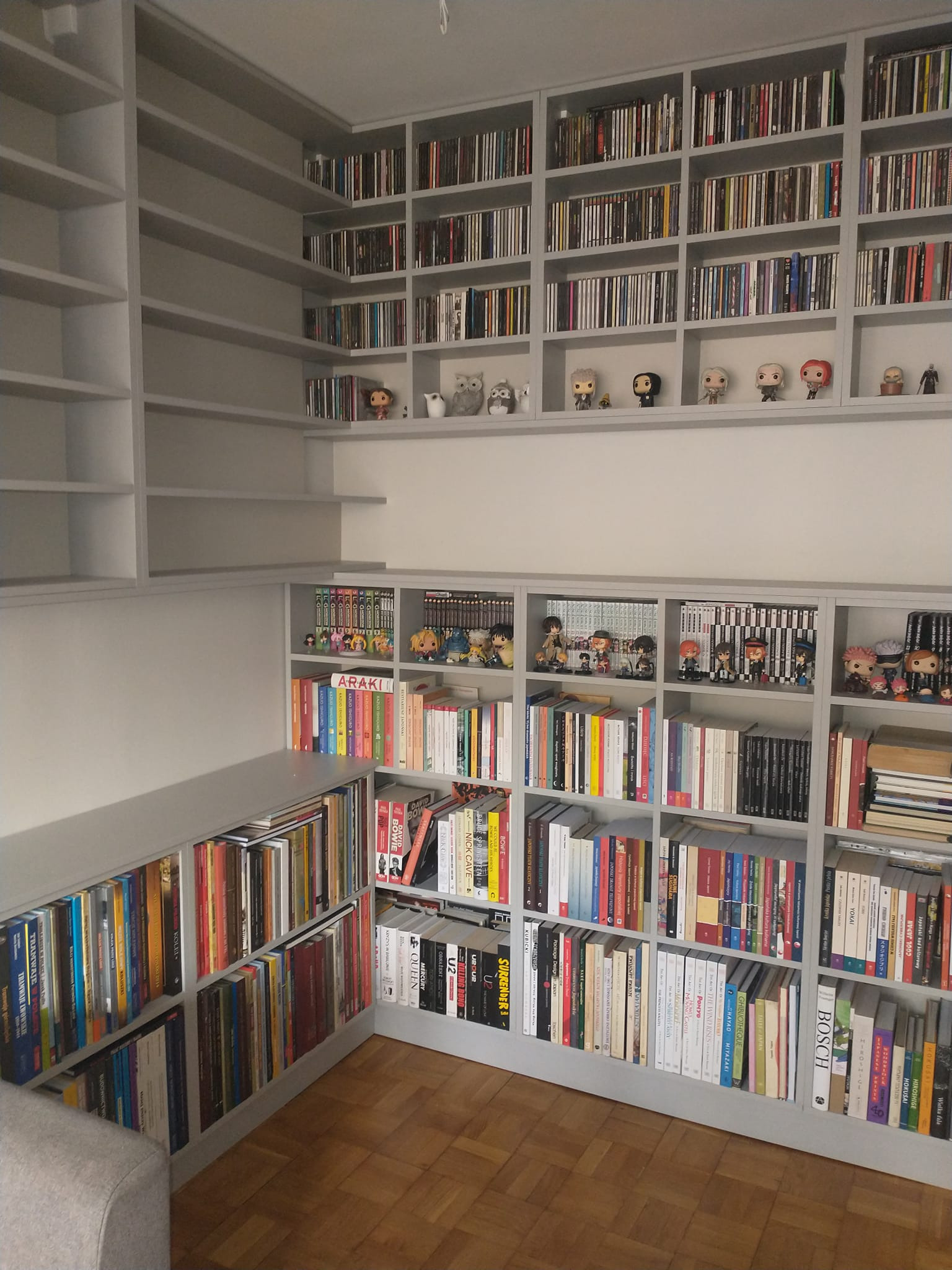 Szara, zabudowana biblioteczka od podłogi do sufitu, wypełniona książkami, płytami CD i figurkami kolekcjonerskimi, z parkietem na podłodze.
