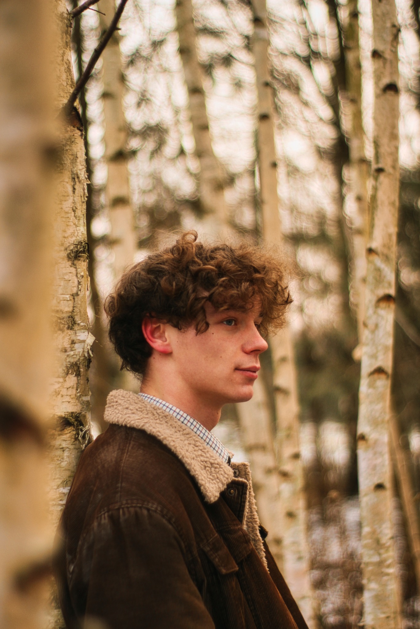 Portret młodego mężczyzny z kręconymi włosami, ubranego w brązową kurtkę z kożuszkiem, stojącego na tle brzozowego lasu w pochmurny dzień.
