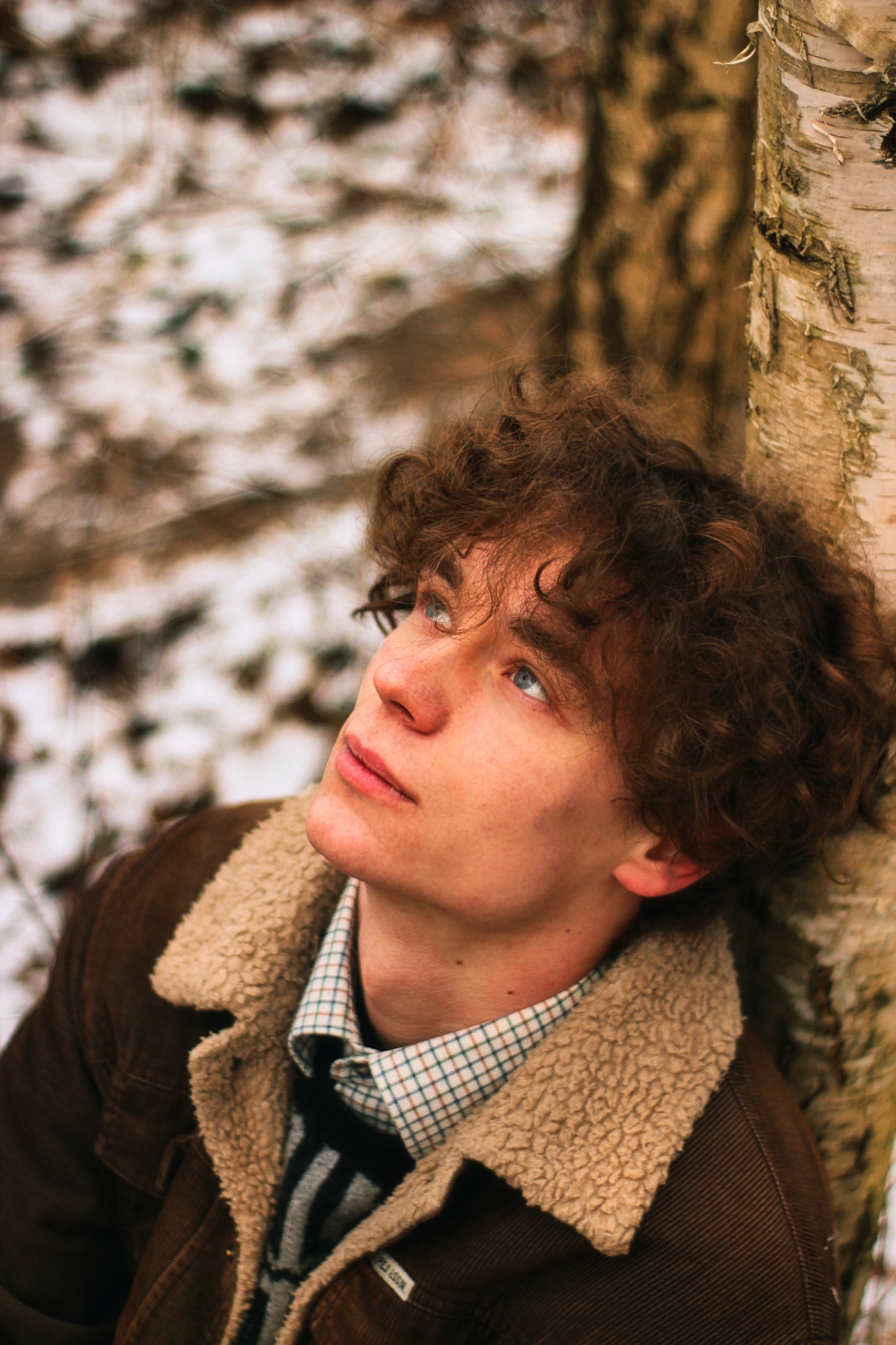 Młody mężczyzna z kręconymi włosami, oparty o korę brzozy, patrzy w górę, ubrany w brązową kurtkę z kożuszkiem i koszulę w kratę.