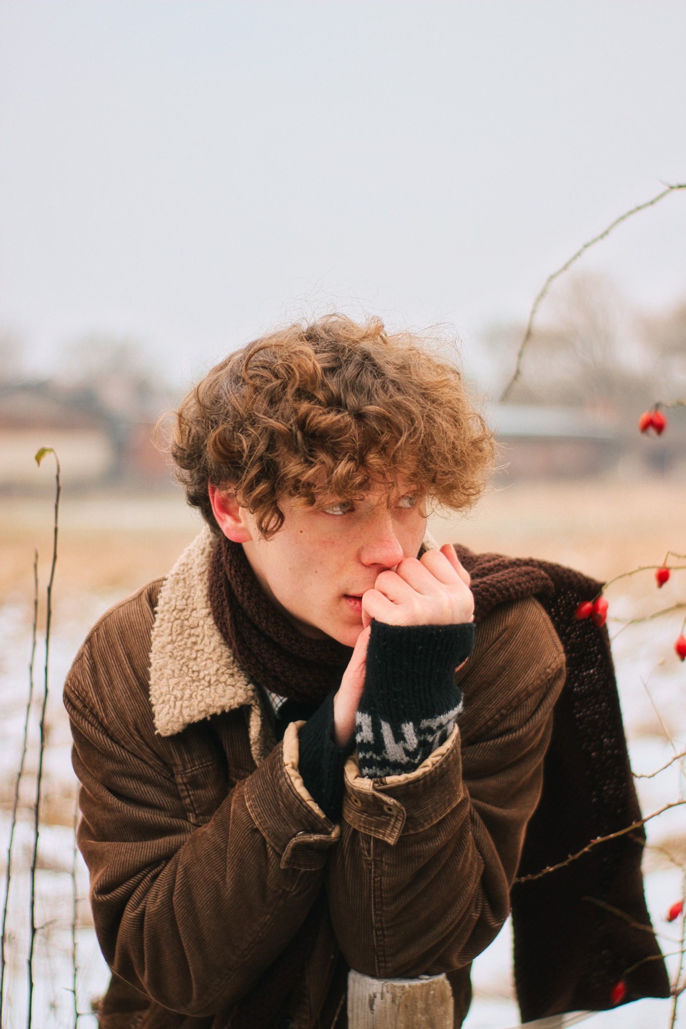 Młody mężczyzna z kręconymi włosami w brązowej kurtce i szaliku, oparty o drewniany słupek, zamyślony wzrok skierowany w bok, na tle zimowego krajobrazu z krzewami dzikiej róży.
