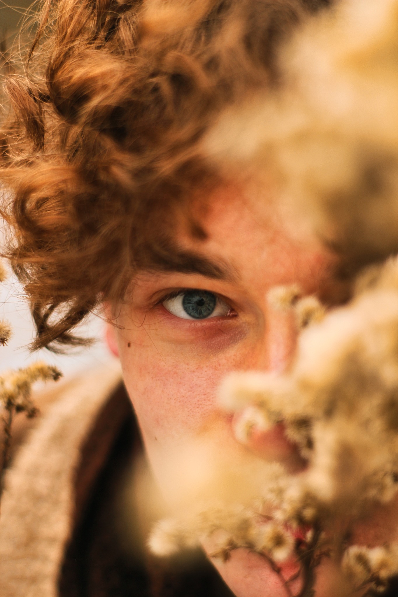 Portret z bliska: Twarz młodej osoby o rudych włosach i niebieskich oczach, częściowo zasłonięta przez suszone kwiaty.