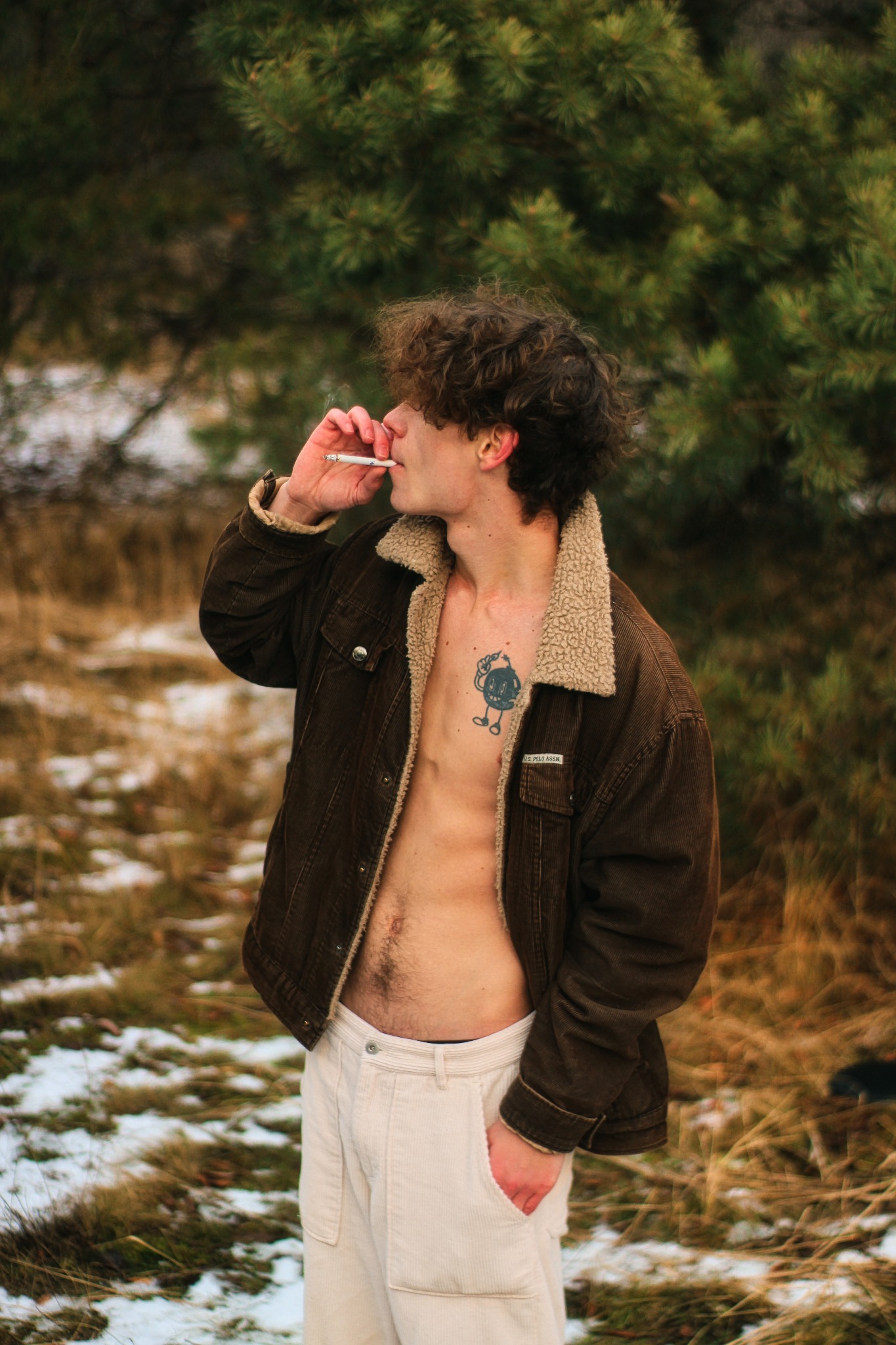 Młody mężczyzna z tatuażem na klatce piersiowej, w rozpiętej kurtce z kożuszkiem i białych spodniach, pali papierosa na tle zimowego krajobrazu w okolicach Łodzi.