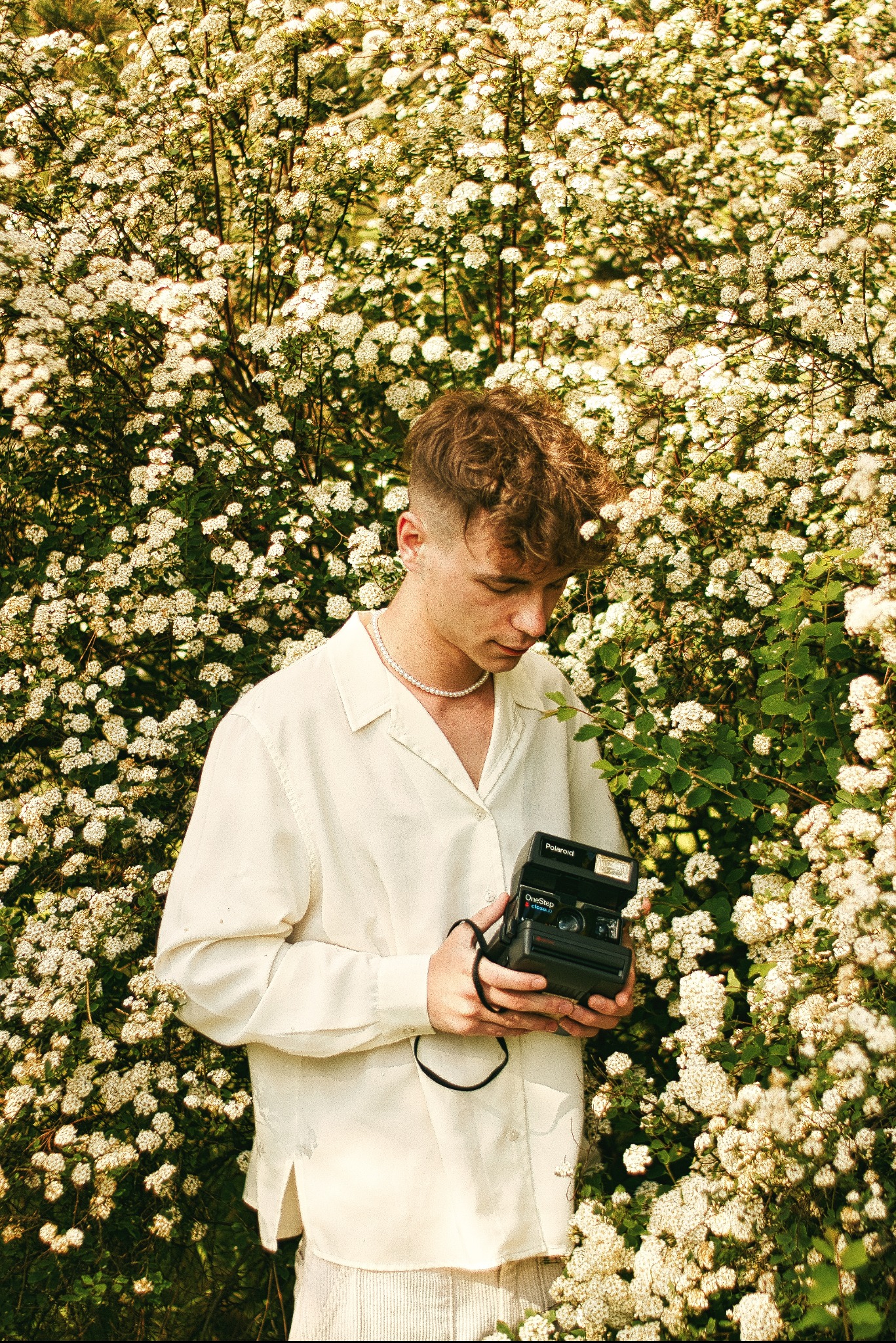Młody mężczyzna w lnianej koszuli i perłach, trzymający aparat Polaroid OneStep w kwitnącym krzewie Spirea.