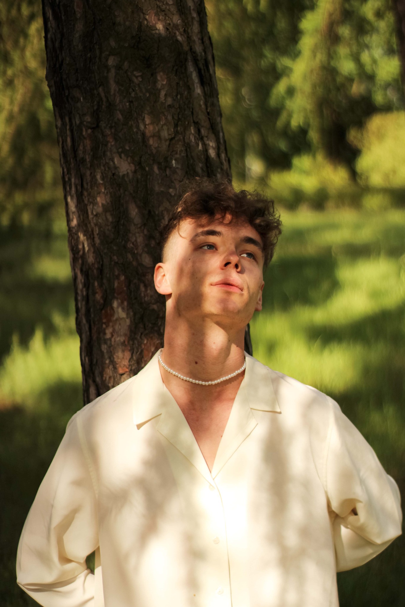 Młody mężczyzna w jasnej koszuli i perłowym naszyjniku opiera się o drzewo w słonecznym lesie, spoglądając w górę.
