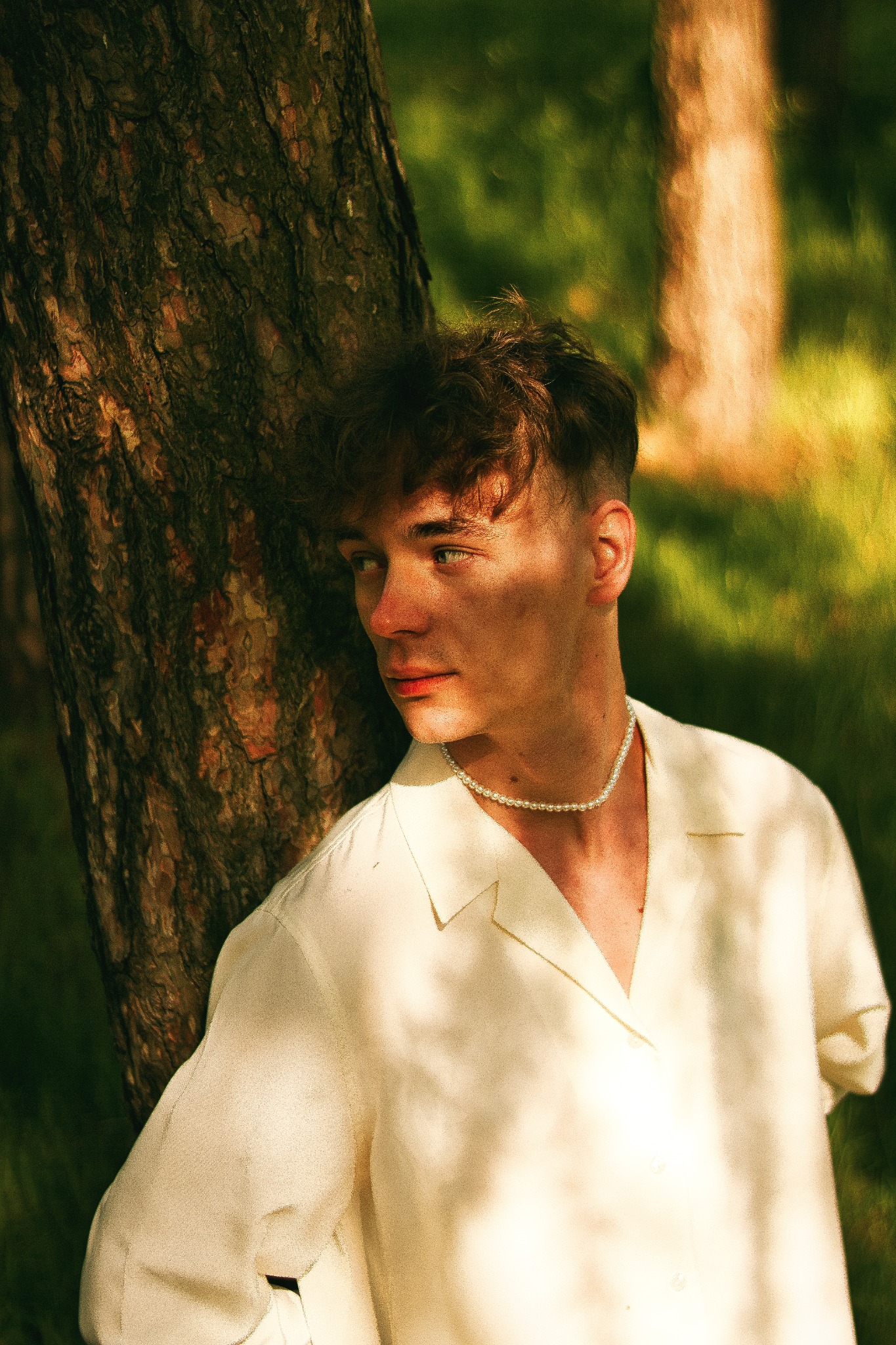 Młody mężczyzna w białej koszuli i perłowym naszyjniku, oparty o drzewo na tle rozmytej zieleni.