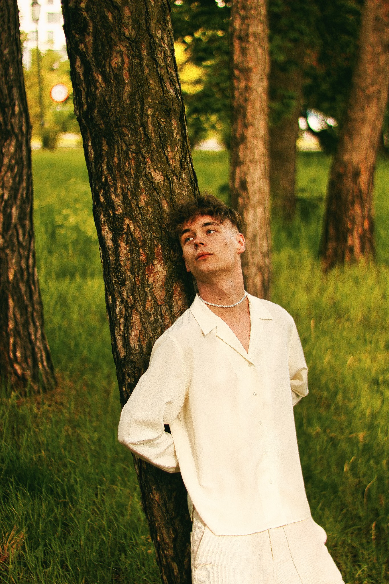 Młody mężczyzna w białej koszuli i spodniach opiera się o drzewo w parku, spoglądając w górę, z zielonym trawiastym tłem i innymi drzewami w tle.